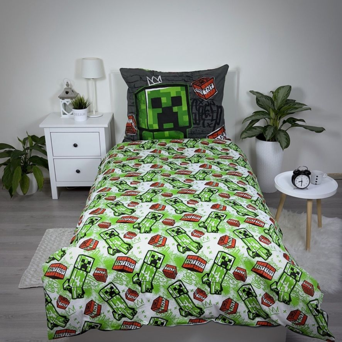 Ein Minecraft-Bettwäsche-Set. Es zeigt ein grünes Creeper- und TNT-Muster auf Bettdecke und Kissenbezug. Nachttisch mit Lampe und Pflanze.