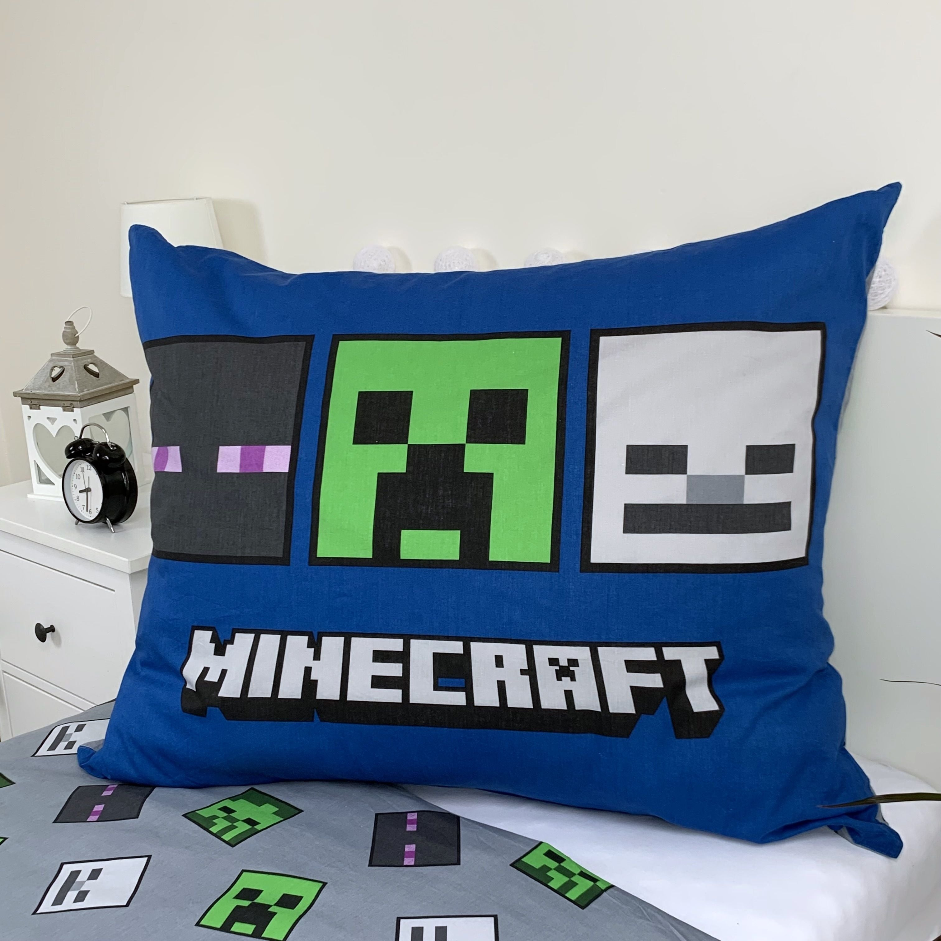 Blaues Minecraft-Kissen auf dem Bett. Es hat Minecraft-Charaktere: Enderman, Creeper und ein Skelett. Nachttisch und Uhr sind sichtbar.