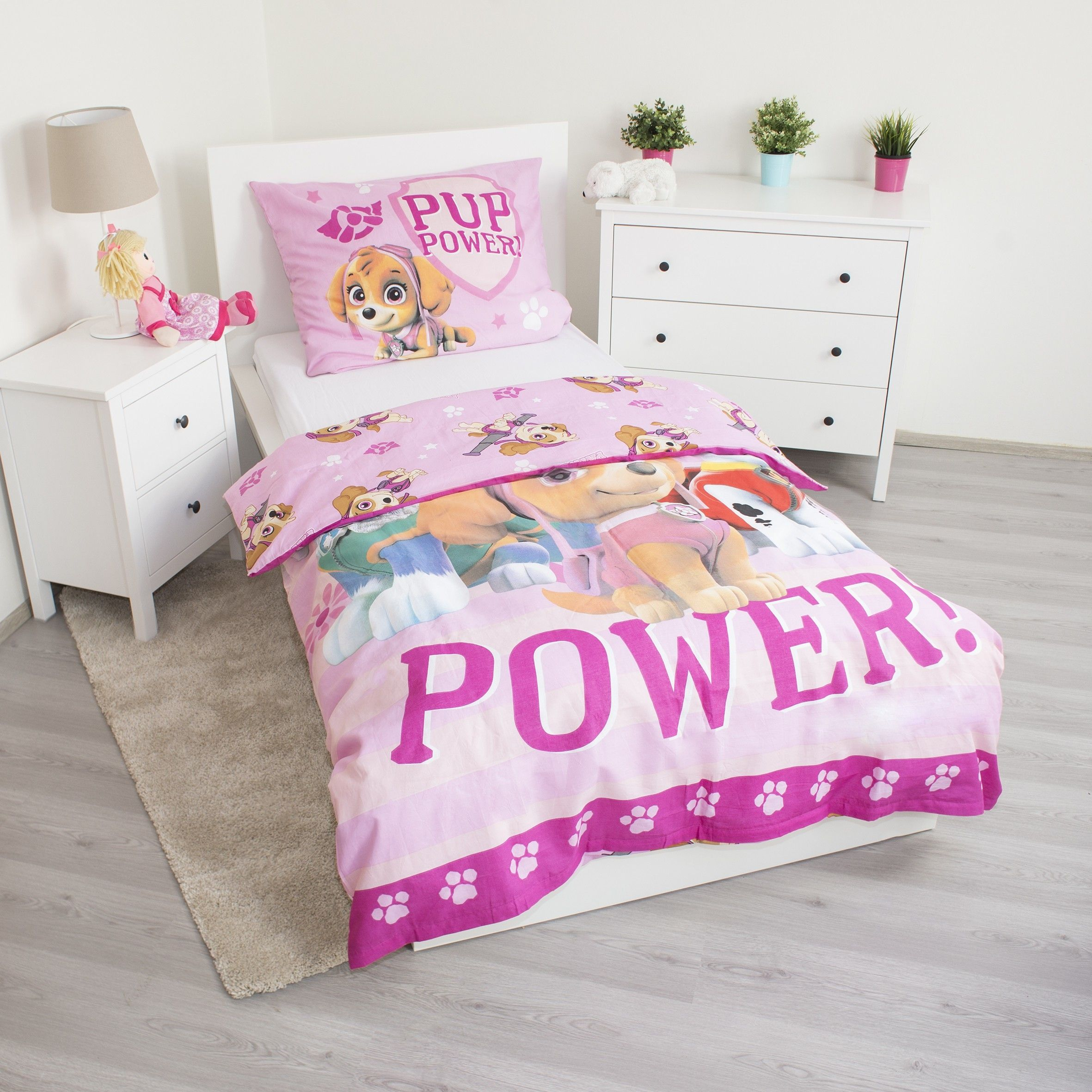 Ein Kinderzimmer mit einem Bett mit Paw Patrol-Bettwäsche. Eine Lampe, eine Puppe und ein Schrank sind zu sehen. Pflanzen und ein Teppich sind vorhanden.