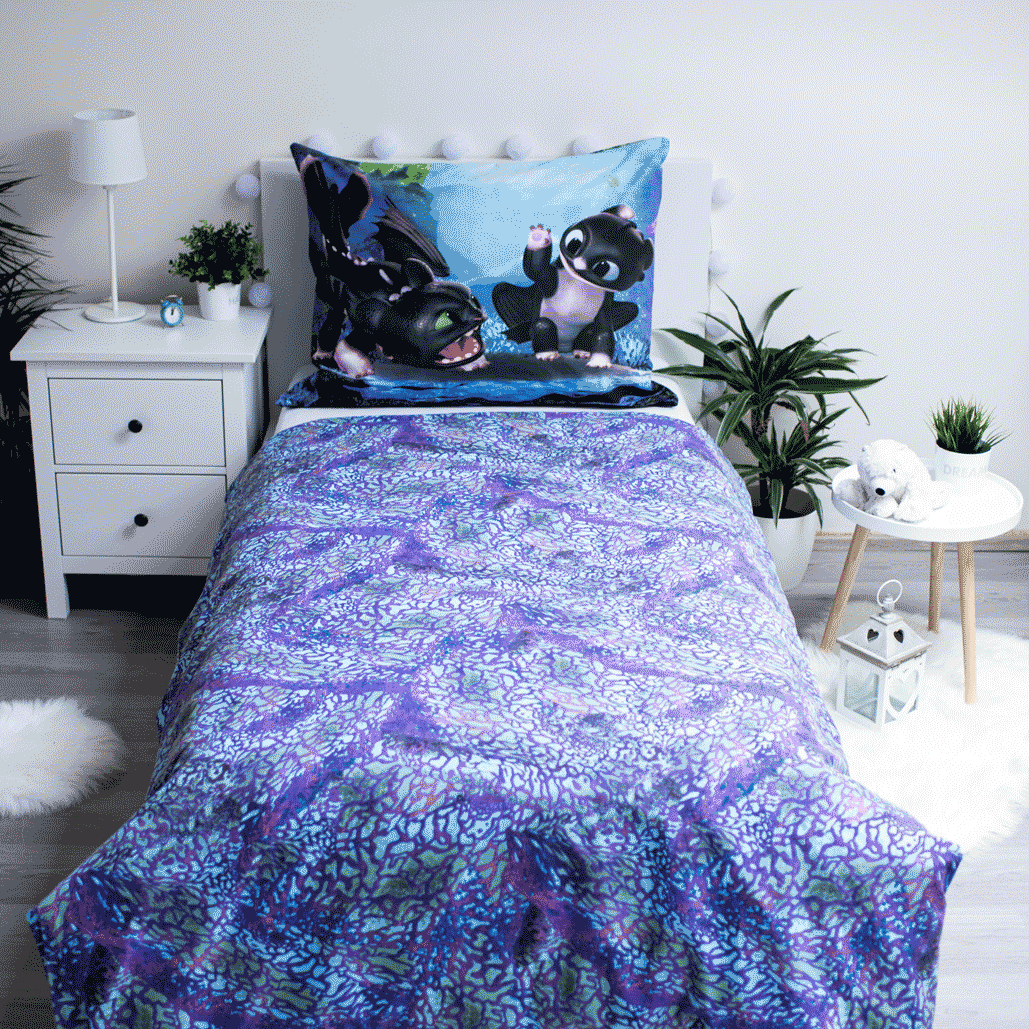 Ein Bett mit einer gemusterten Bettdecke und einem Kissen mit Zeichentrickdrachen. Ein Nachttisch mit Lampe und Pflanzen. Ein kleiner Tisch.
