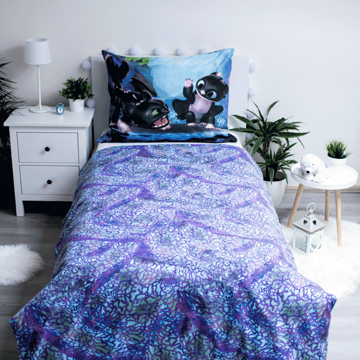 Bett mit lilafarbenem Muster-Bettbezug. Kissen zeigt Ohnezahn-Drachen. Pflanzen und weiße Möbel umgeben das Bett.