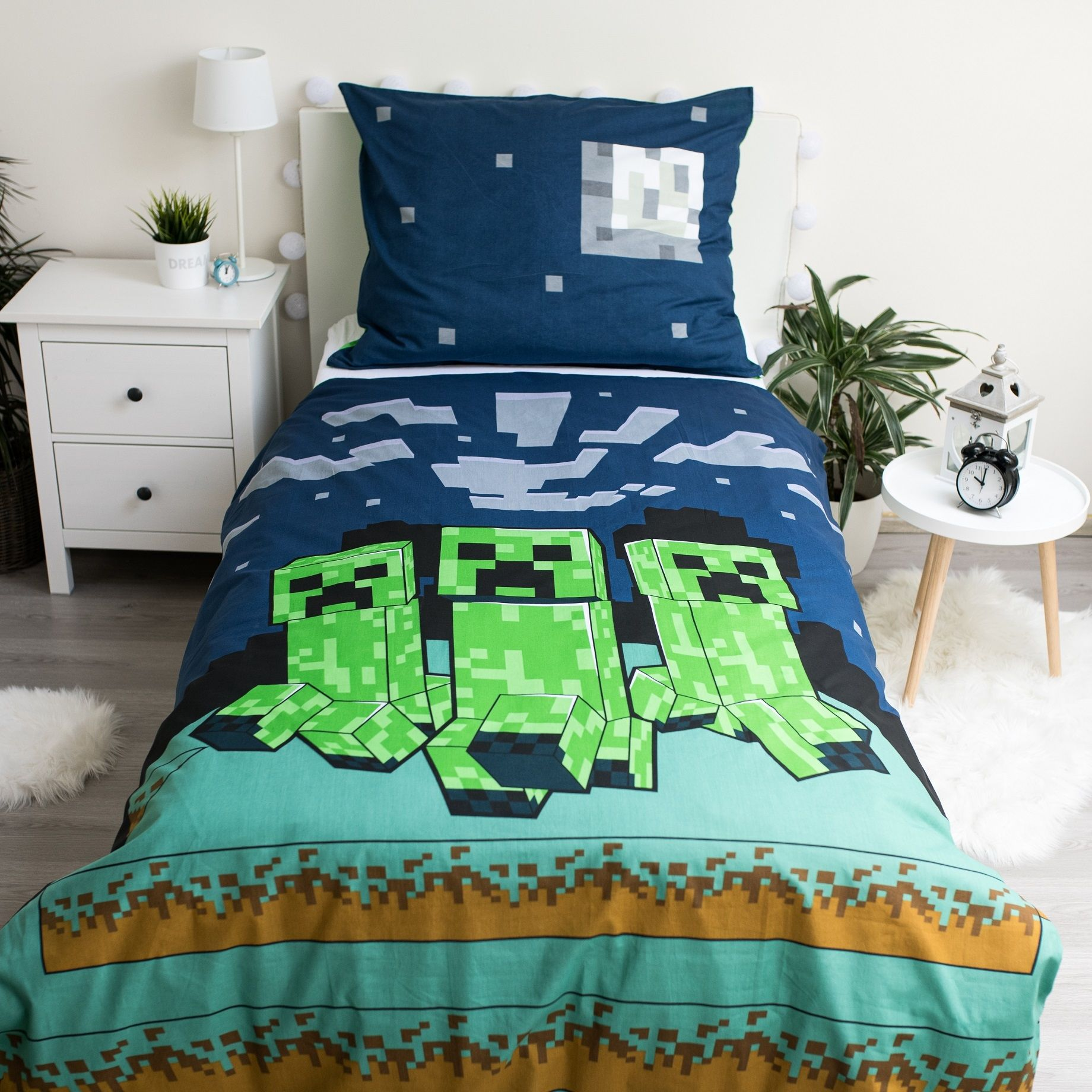 Bett mit Bettwäsche im Minecraft-Design mit grünen Creeper-Charakteren. Weiße Nachttische, Pflanzen und ein kleiner Tisch vervollständigen den Raum.