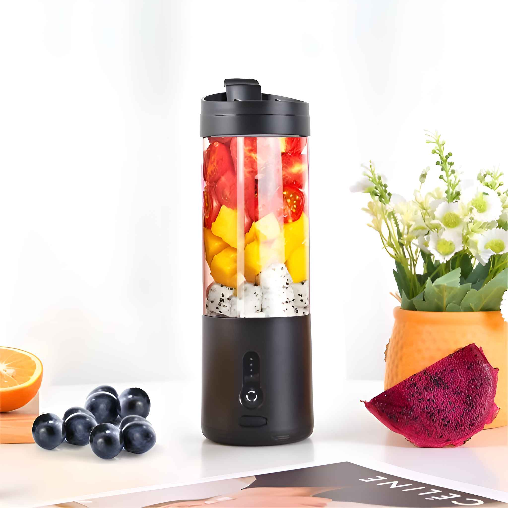 Ein schwarzer Mixer mit Früchten steht auf einer weißen Oberfläche. Trauben, Orangenscheibe, Blumen und ein Drachenfruchtstück sind in der Nähe.