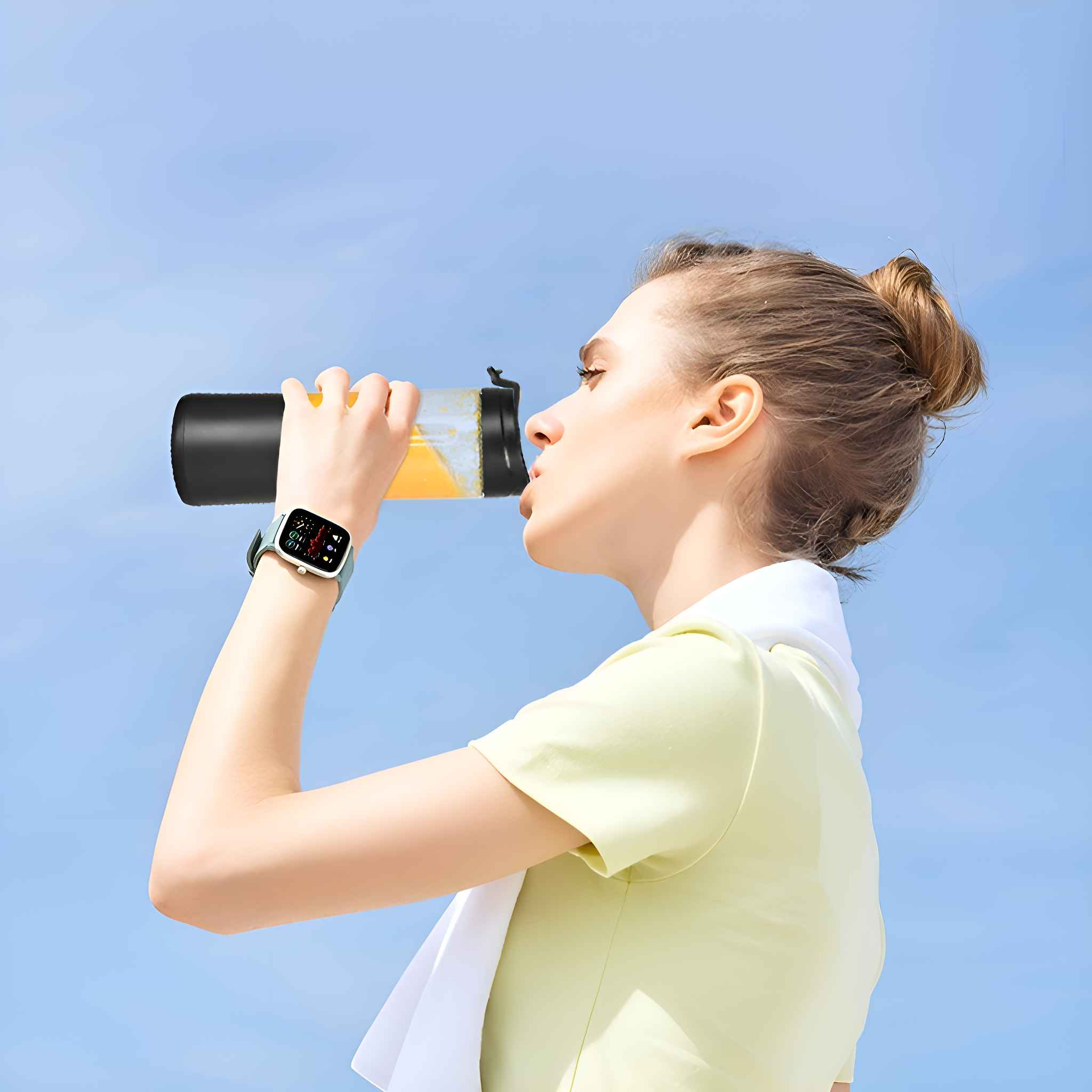 Eine Frau trinkt aus einer Sportflasche. Sie trägt eine Uhr, ein helles Hemd und ein weißes Handtuch. Der Hintergrund ist ein klarer blauer Himmel.