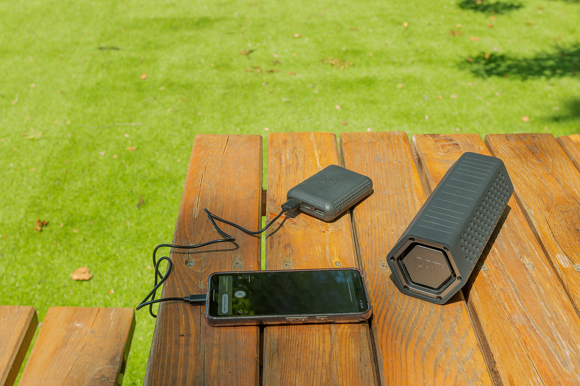 Un smartphone, un banco de energía y un altavoz sobre una mesa de madera. Los cables conectan los dispositivos. El fondo es hierba verde.