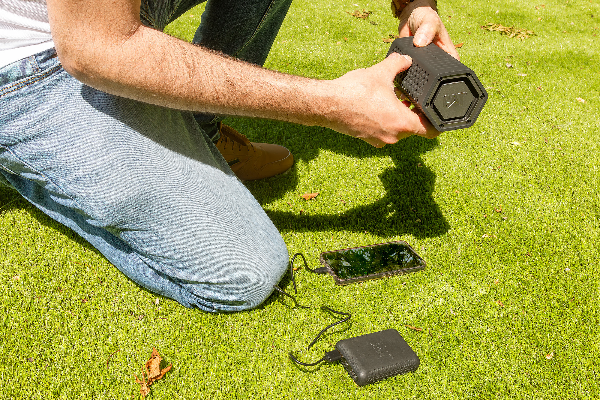 Persona arrodillada en la hierba, sosteniendo un altavoz negro. Un teléfono y un banco de energía están conectados. La escena es al aire libre en un día soleado.