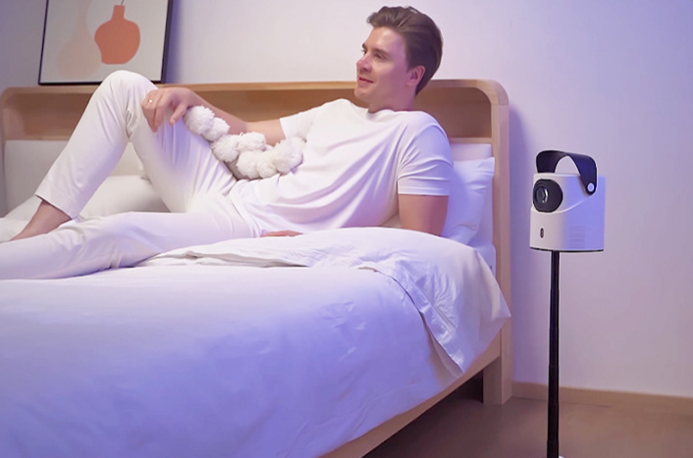 Mann in weißer Kleidung liegt auf einem Bett und hält ein weißes, flauschiges Objekt. Ein Projektor steht daneben.