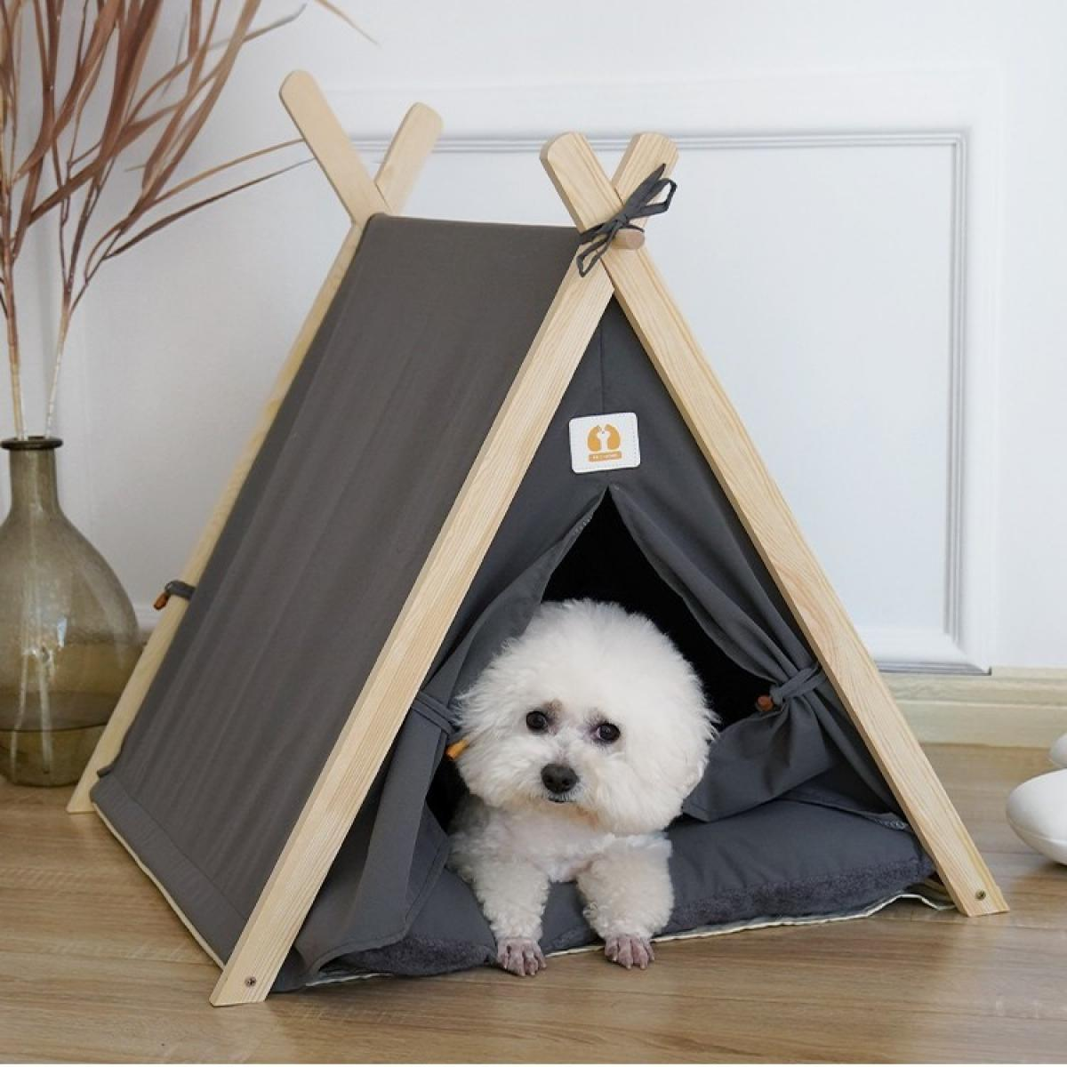 Ein weißer, flauschiger Hund sitzt in einem grau gefärbten, zeltförmigen Hundebett. Das Zelt hat einen Holzrahmen und steht drinnen.