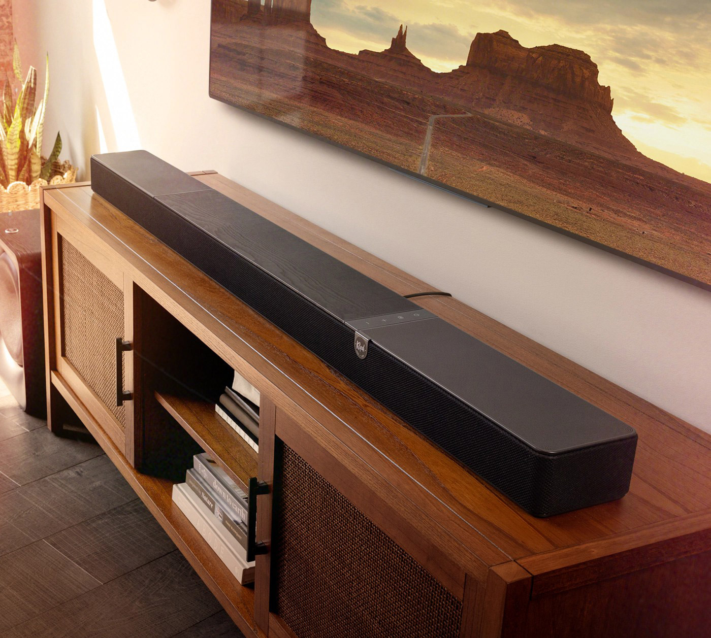 Eine schwarze Soundbar liegt auf einem Holzschrank. Darüber hängt ein Fernseher mit Landschaftsbild. Pflanzen und Bücher sind ebenfalls sichtbar.