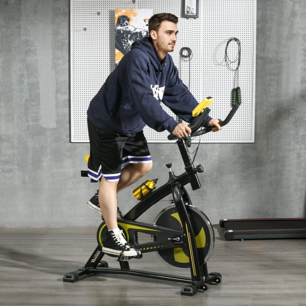 Ein Mann in einem blauen Kapuzenpullover und Shorts trainiert auf einem Heimtrainer. Das Fahrrad ist schwarz und gelb. Eine Wasserflasche ist befestigt.