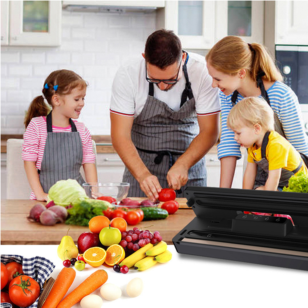 Familie in der Küche bereitet Essen zu. Mann schneidet Tomaten, Kinder und Frau schauen zu. Obst und Gemüse auf dem Tisch. Vakuumierer im Vordergrund.