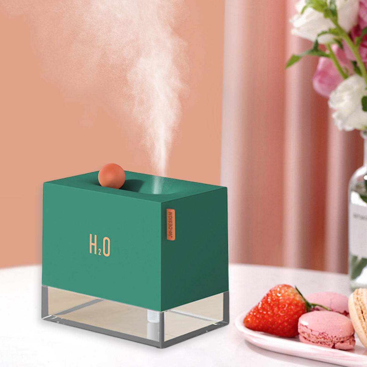 Ein grüner Luftbefeuchter mit H2O darauf. Eine Erdbeere und Makronen befinden sich auf einem Teller. Blumen sind im Hintergrund.