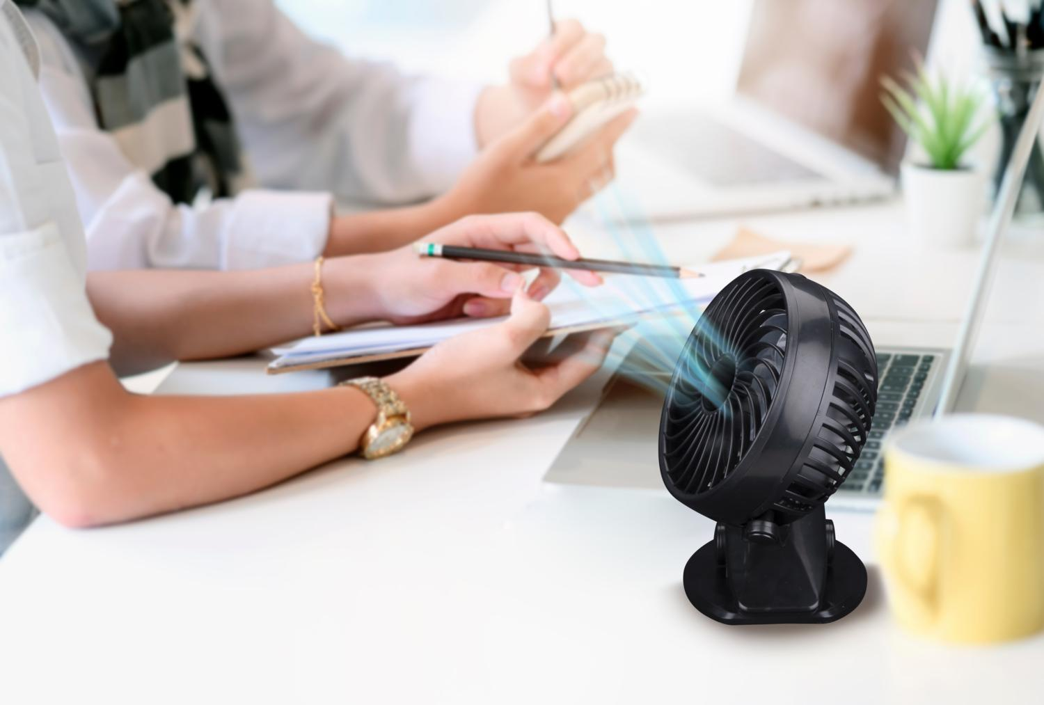 Un ventilateur de bureau noir souffle de l'air vers des personnes travaillant à un bureau avec un ordinateur portable. Tasse jaune et pot de plante sont également sur le bureau.