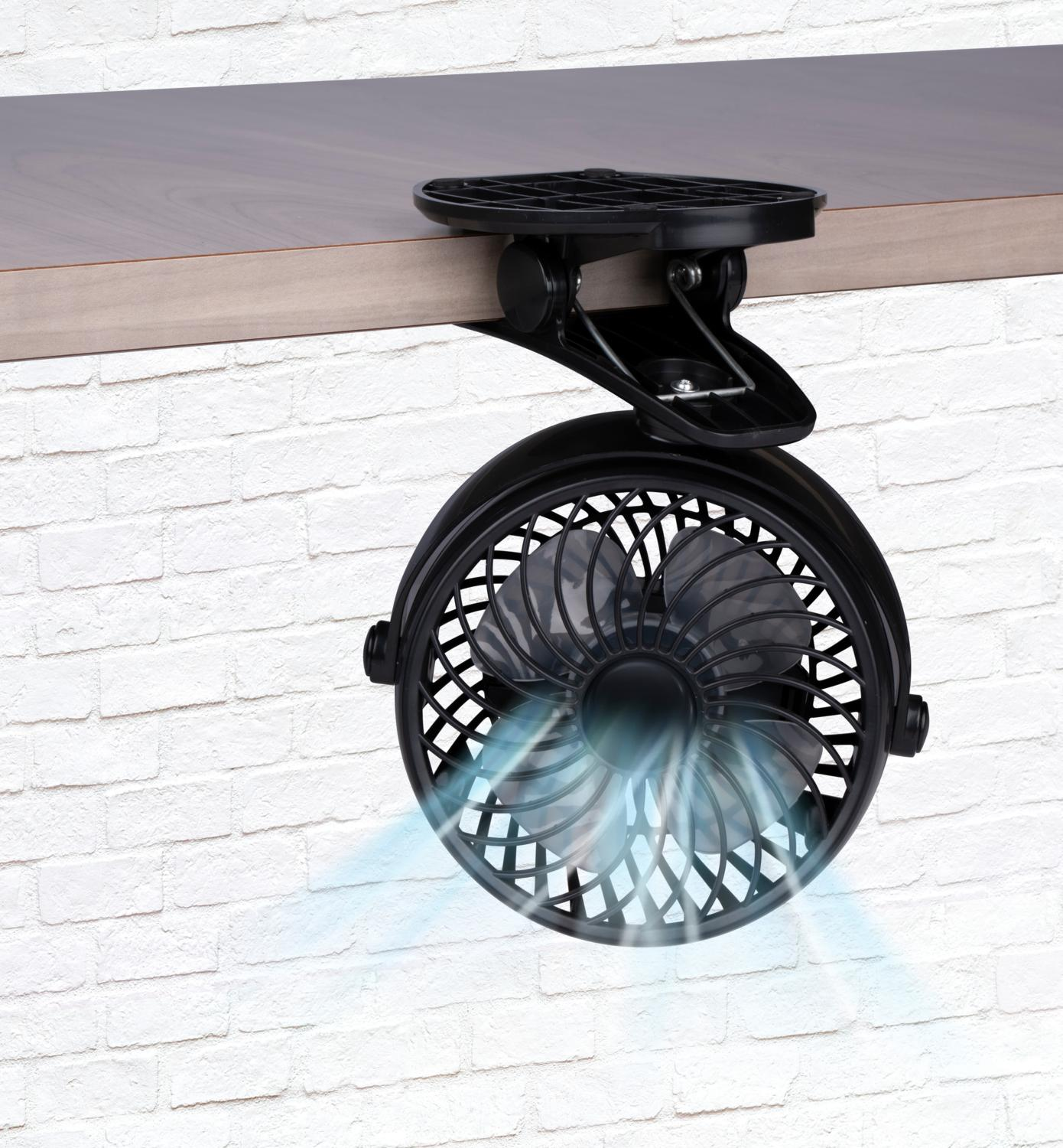 Un ventilateur noir à pince est fixé à une surface en bois, soufflant de l'air contre un mur de briques blanches.