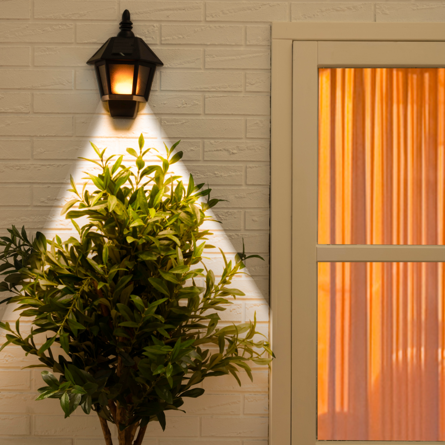 A lit outdoor lamp on a brick wall, illuminating a green plant. To the right, a window with orange curtains is visible.