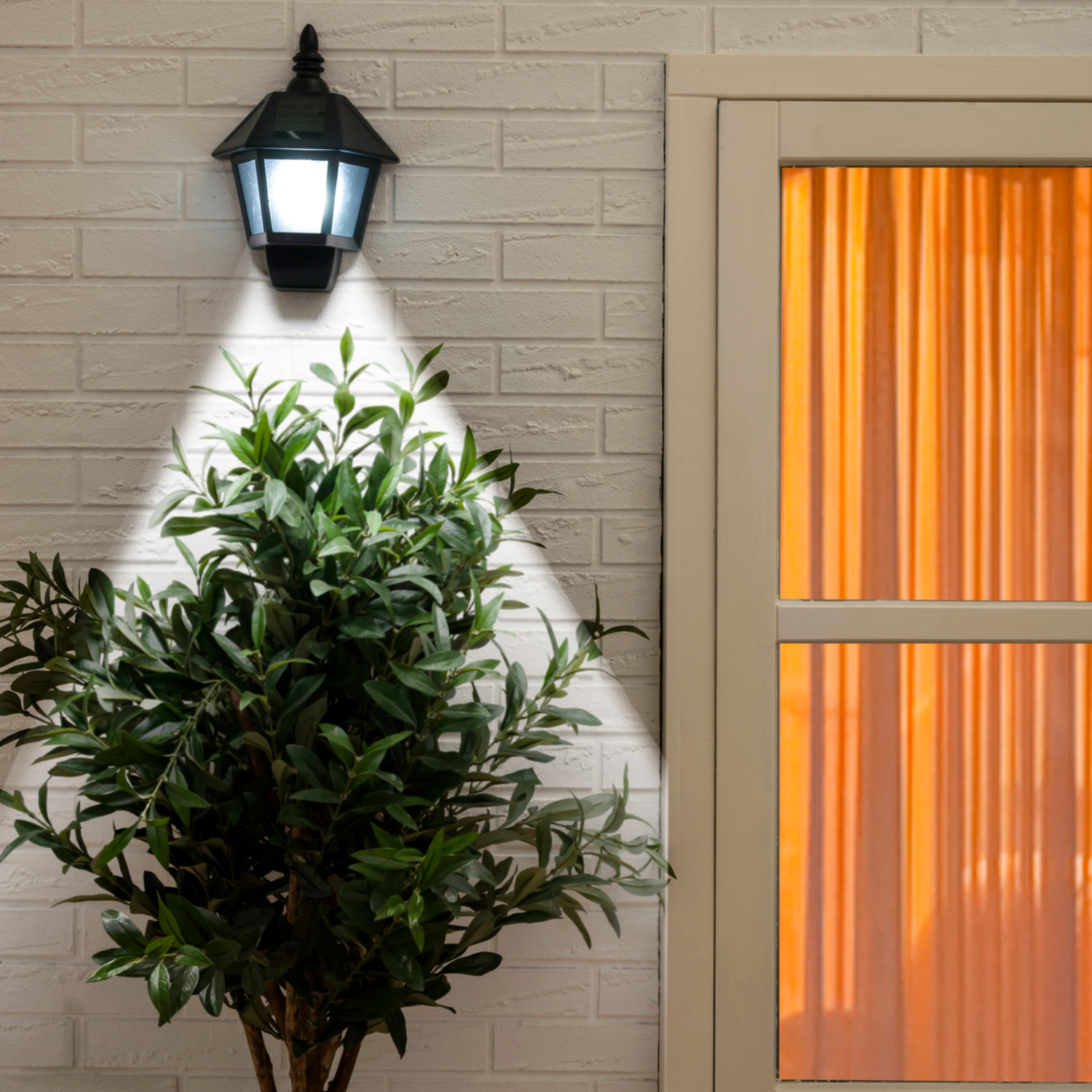 A lit black lamp on a white brick wall. A green bush is in front of the wall, and a window with orange curtains is on the right.
