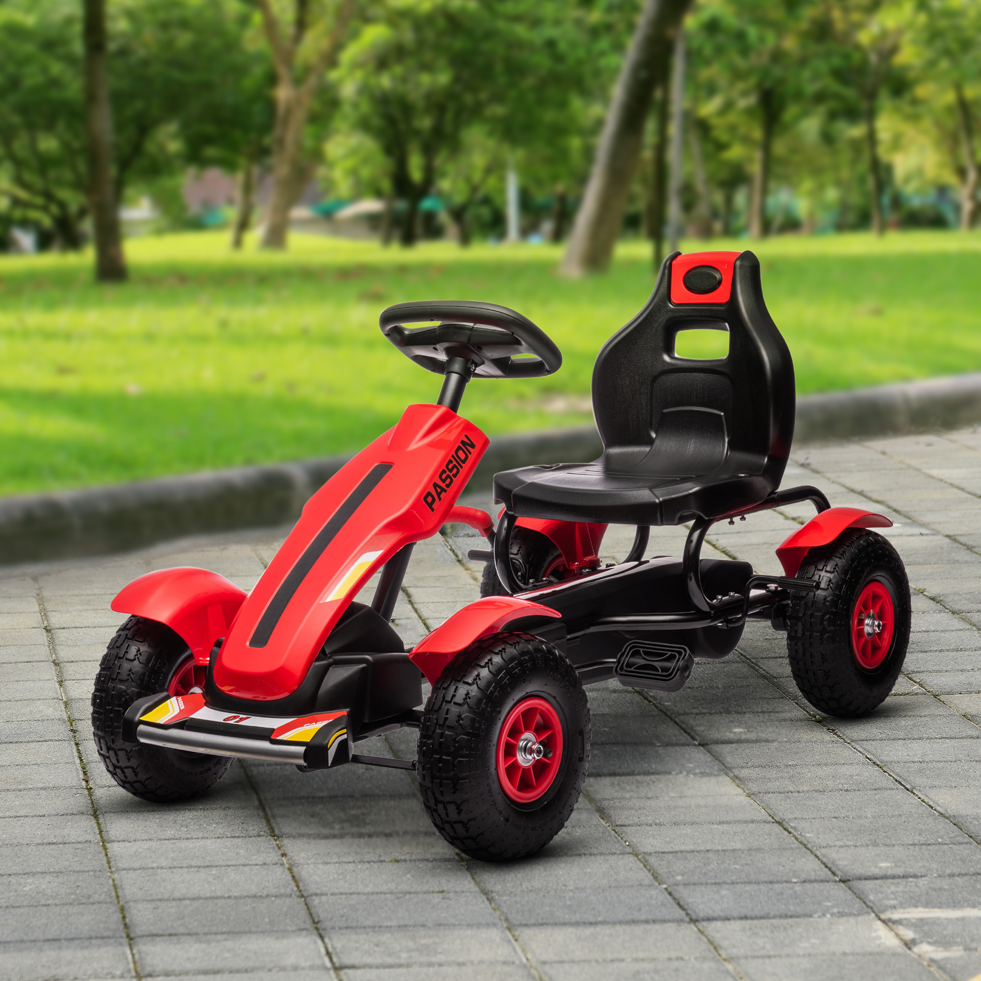 Go-kart a pedali rosso e nero su una superficie pavimentata grigia. Lo sfondo è un parco verde con alberi ed erba.
