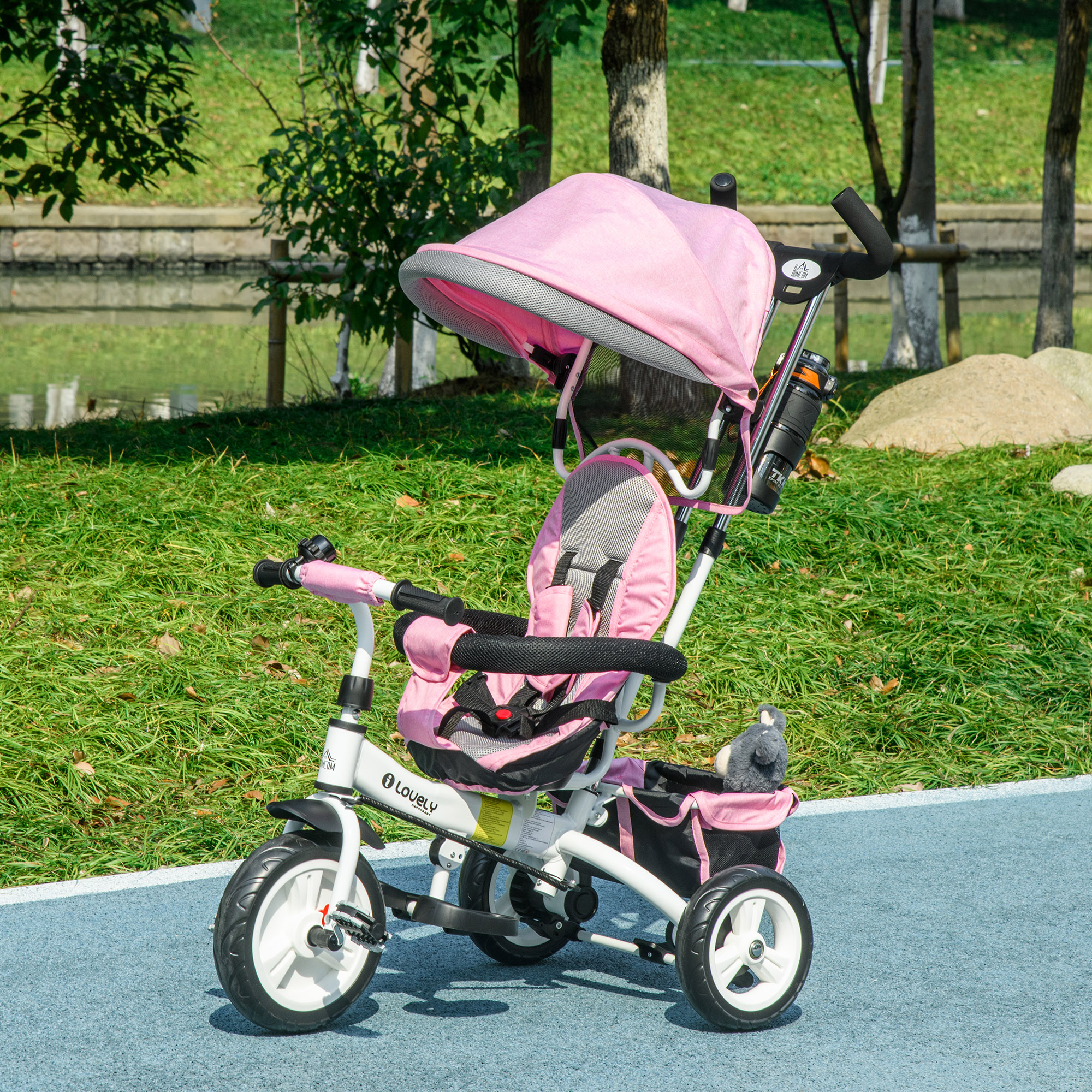 Un triciclo rosa e bianco con parasole si trova su una superficie blu, erba e alberi sullo sfondo.