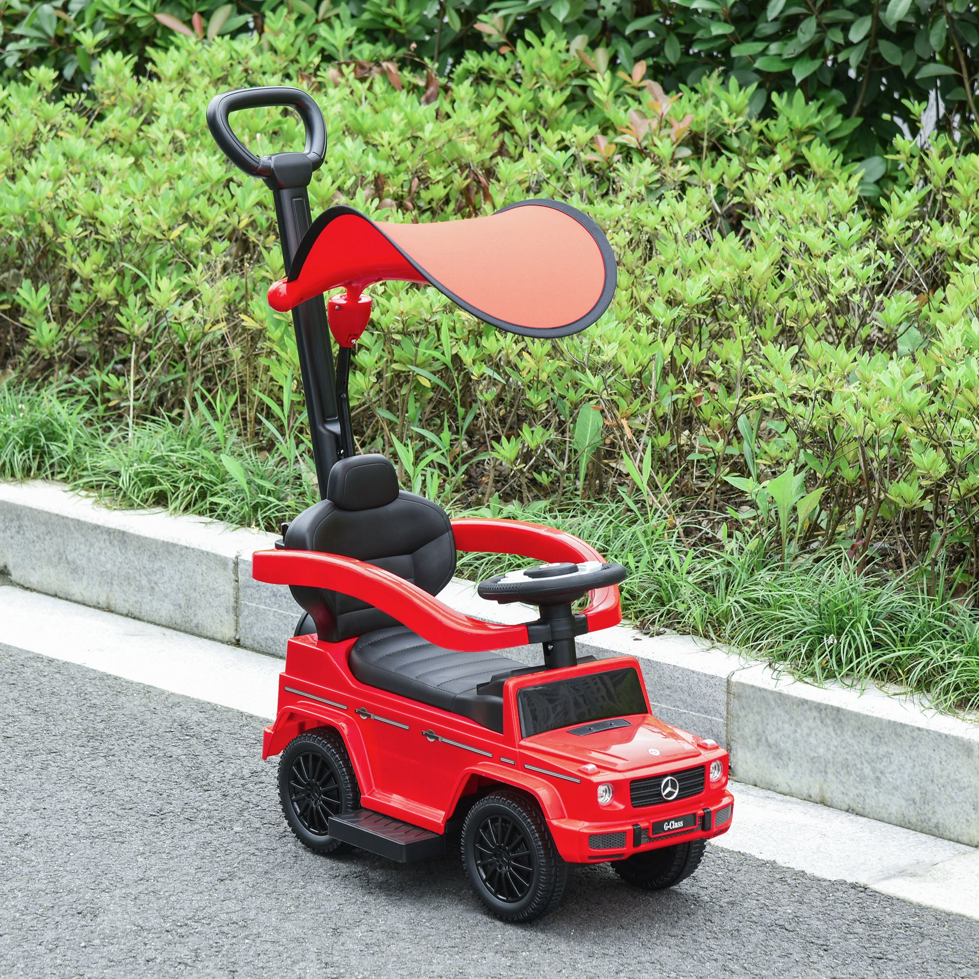 Un'auto giocattolo rossa per bambini con sedile nero e parasole rosso, parcheggiata su un marciapiede grigio. Cespugli verdi sullo sfondo.