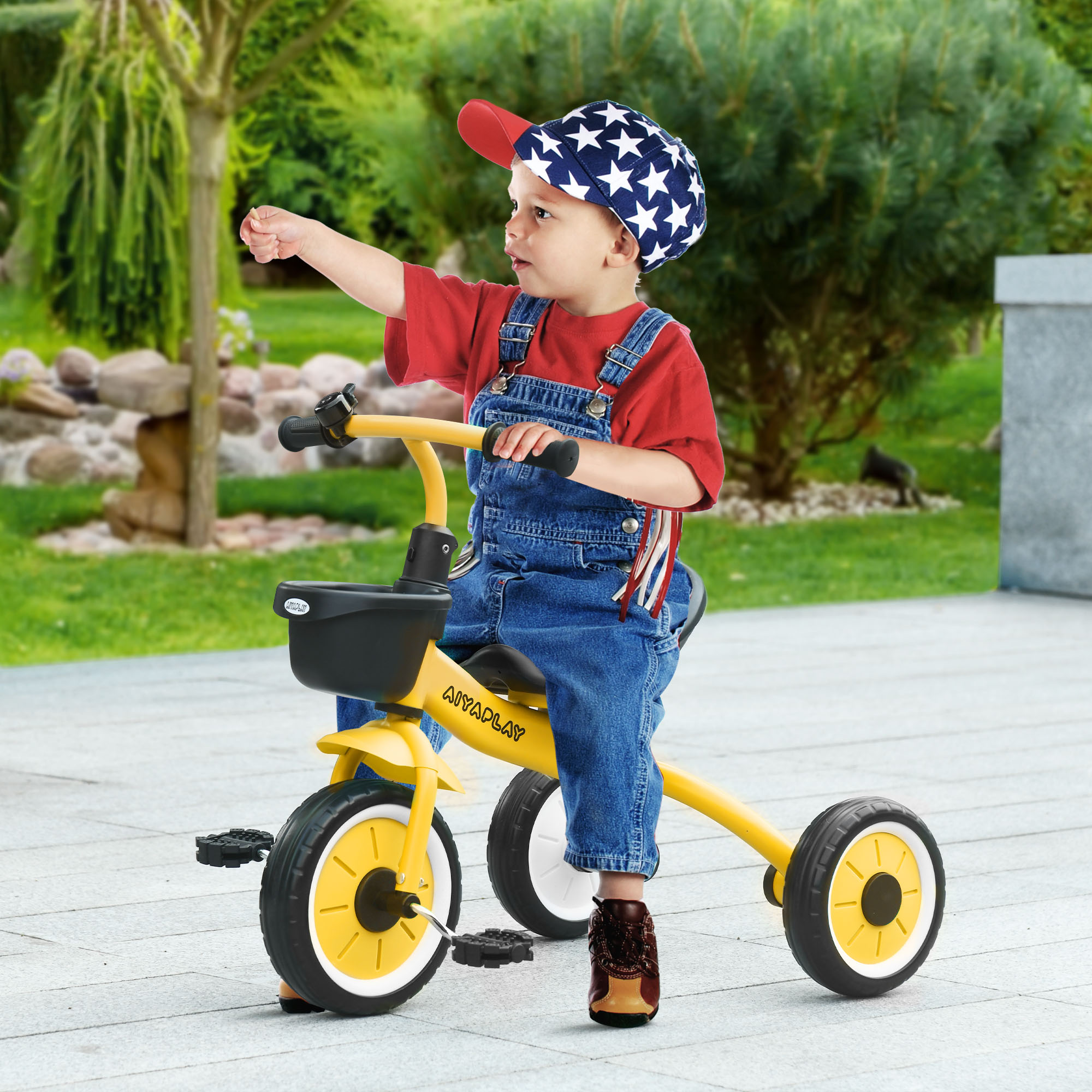 Un bambino con un cappello a stelle cavalca un triciclo giallo, indicando. Sfondo con verde e sentiero di pietra.