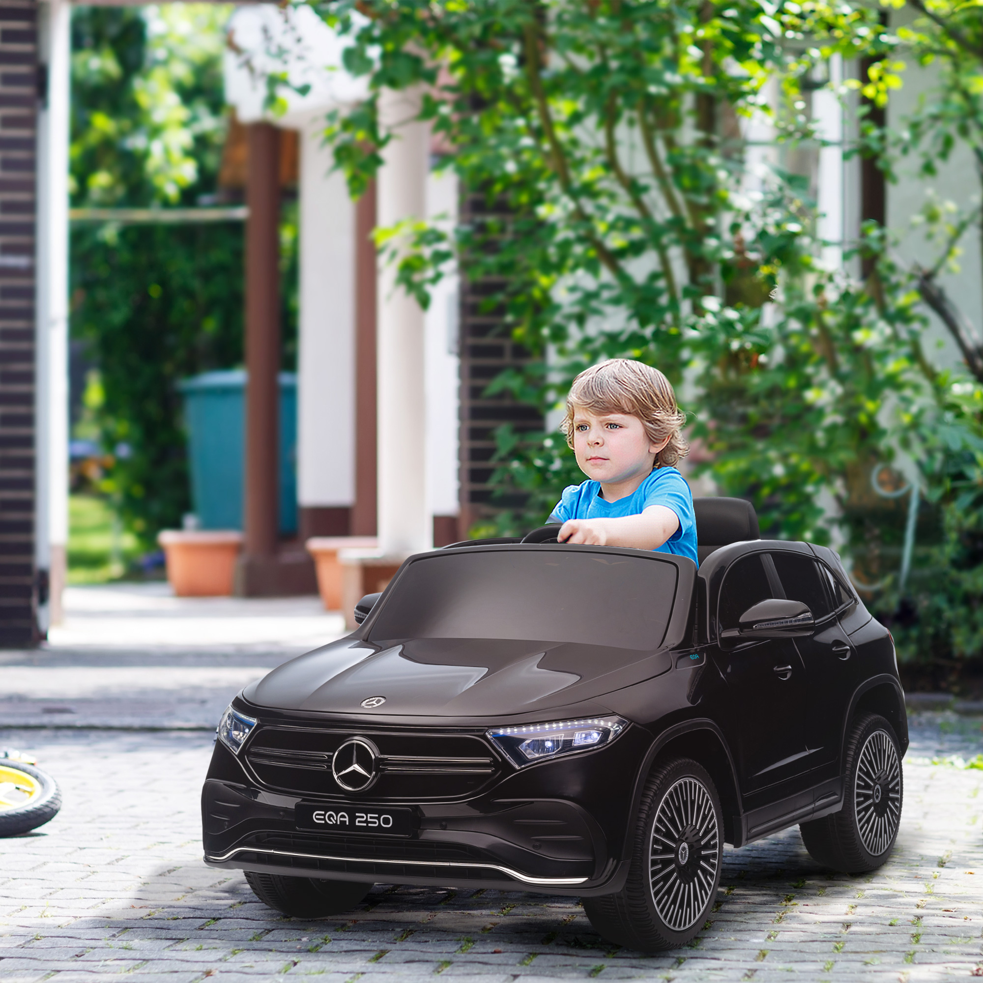 Un bambino in un'auto giocattolo nera. L'auto è una Mercedes EQA 250. Lo sfondo presenta una casa e vegetazione.