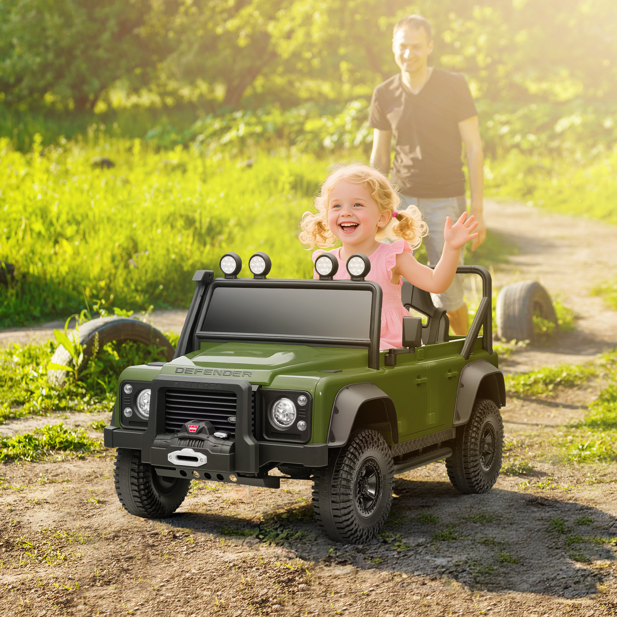 Una bambina saluta da un'auto giocattolo verde. Un uomo cammina dietro. Lo sfondo è un campo verde e soleggiato.