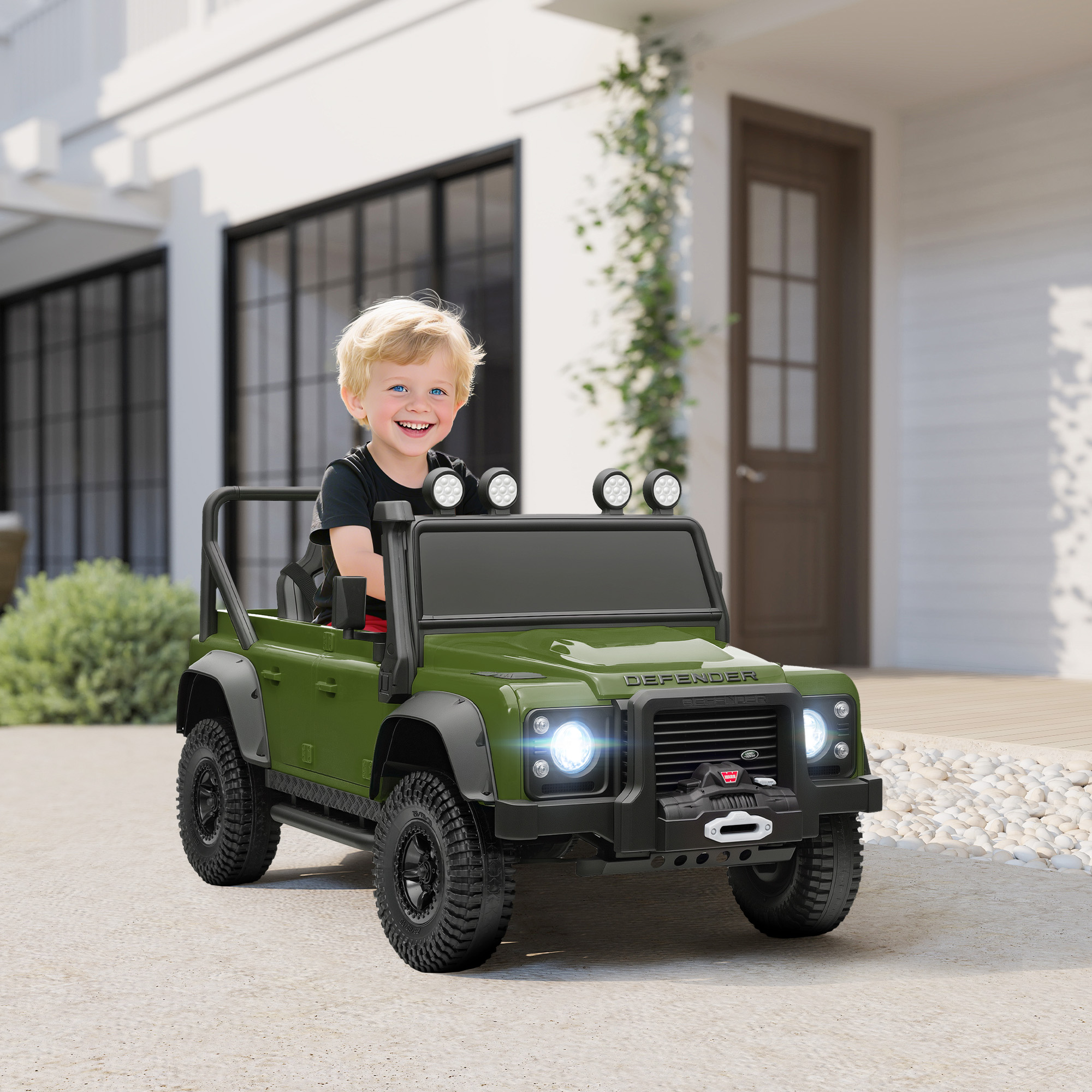 Un bambino sorridente è seduto in un'auto giocattolo verde. È un modello Defender. Lo sfondo mostra una casa e alcuni cespugli.