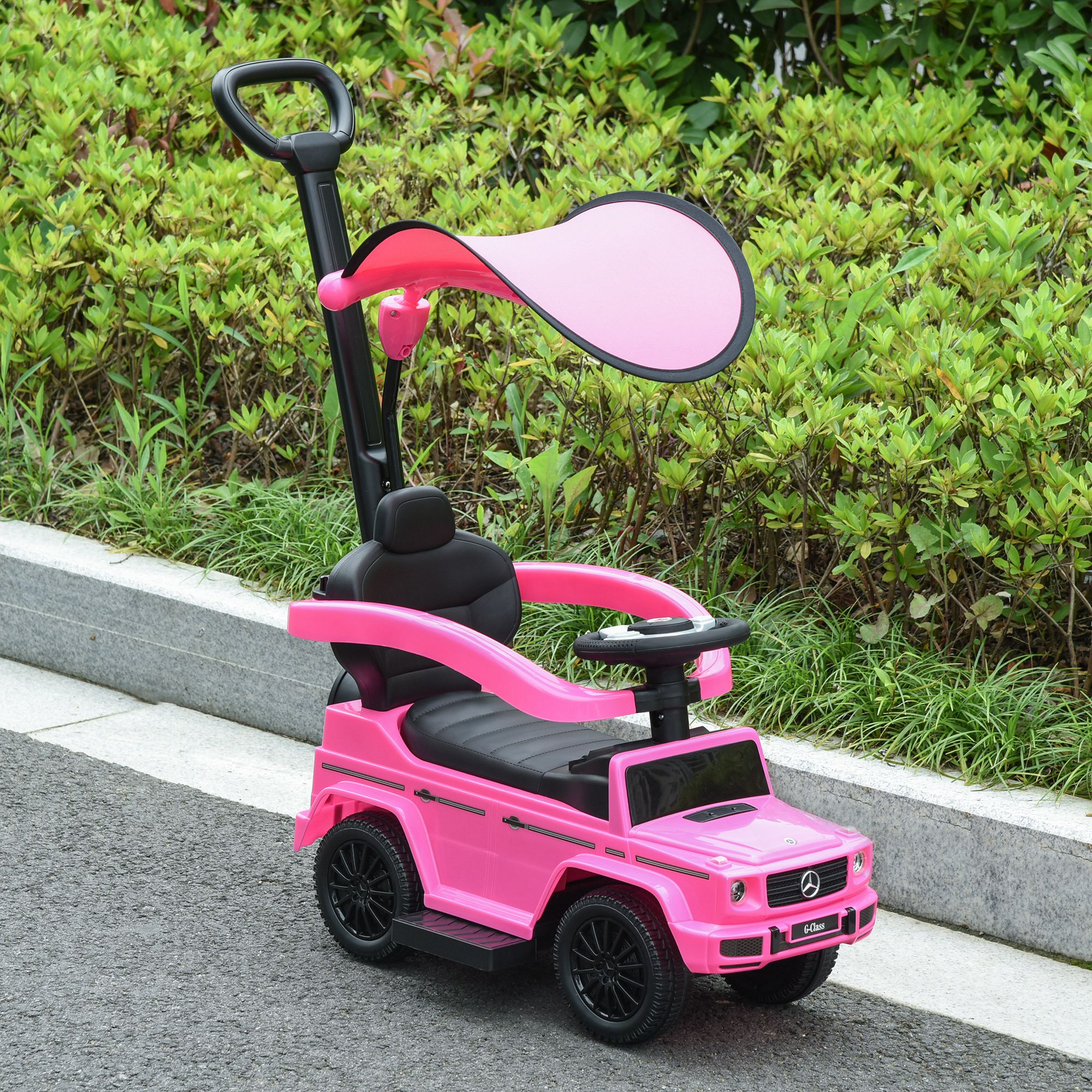 Un'auto giocattolo rosa con ruote nere e tettuccio è sull'asfalto. Cespugli verdi e un bordo di pietra sullo sfondo.