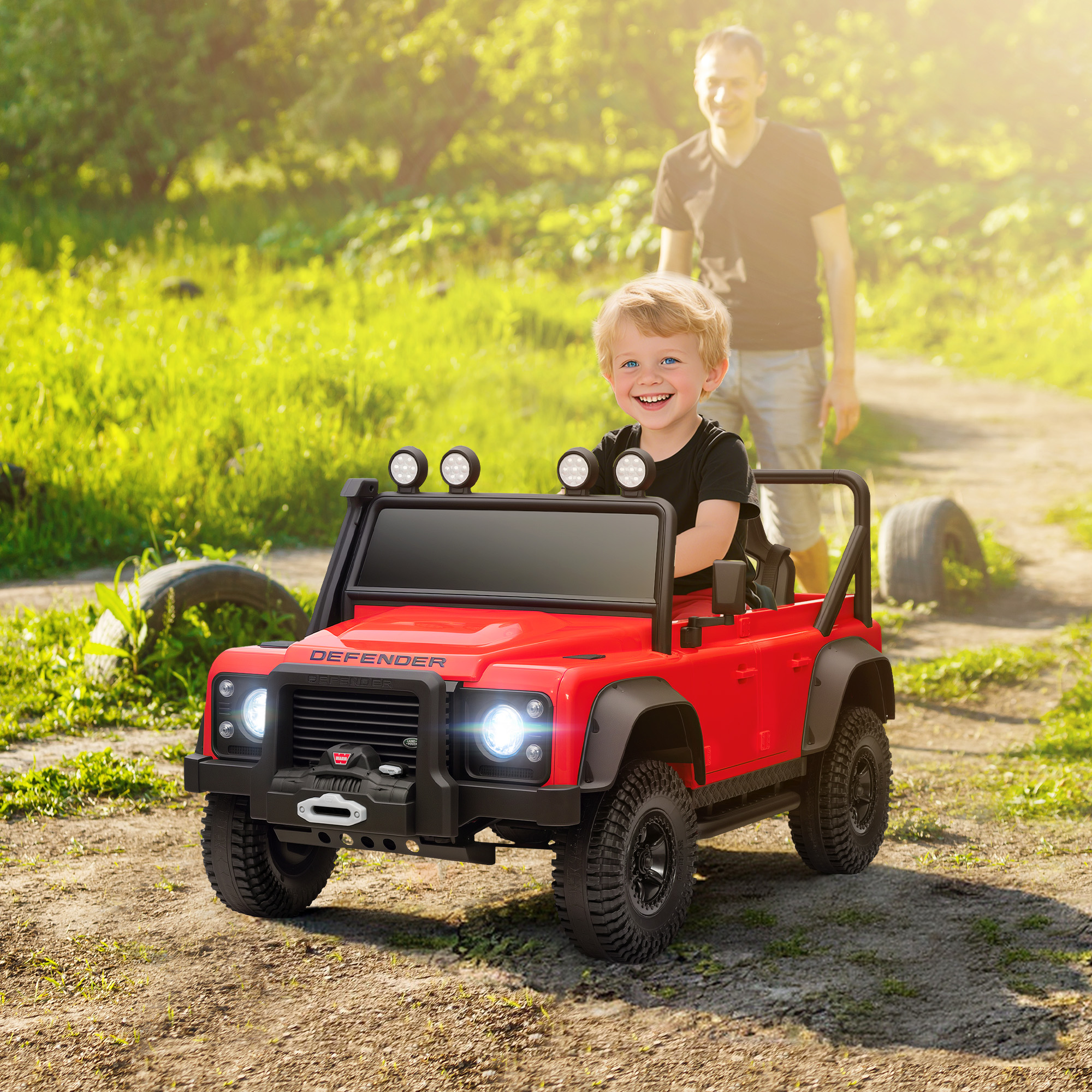 Un niño en un coche de juguete rojo sonríe. Un hombre camina detrás. El coche está en un camino de tierra, rodeado de hierba y árboles.