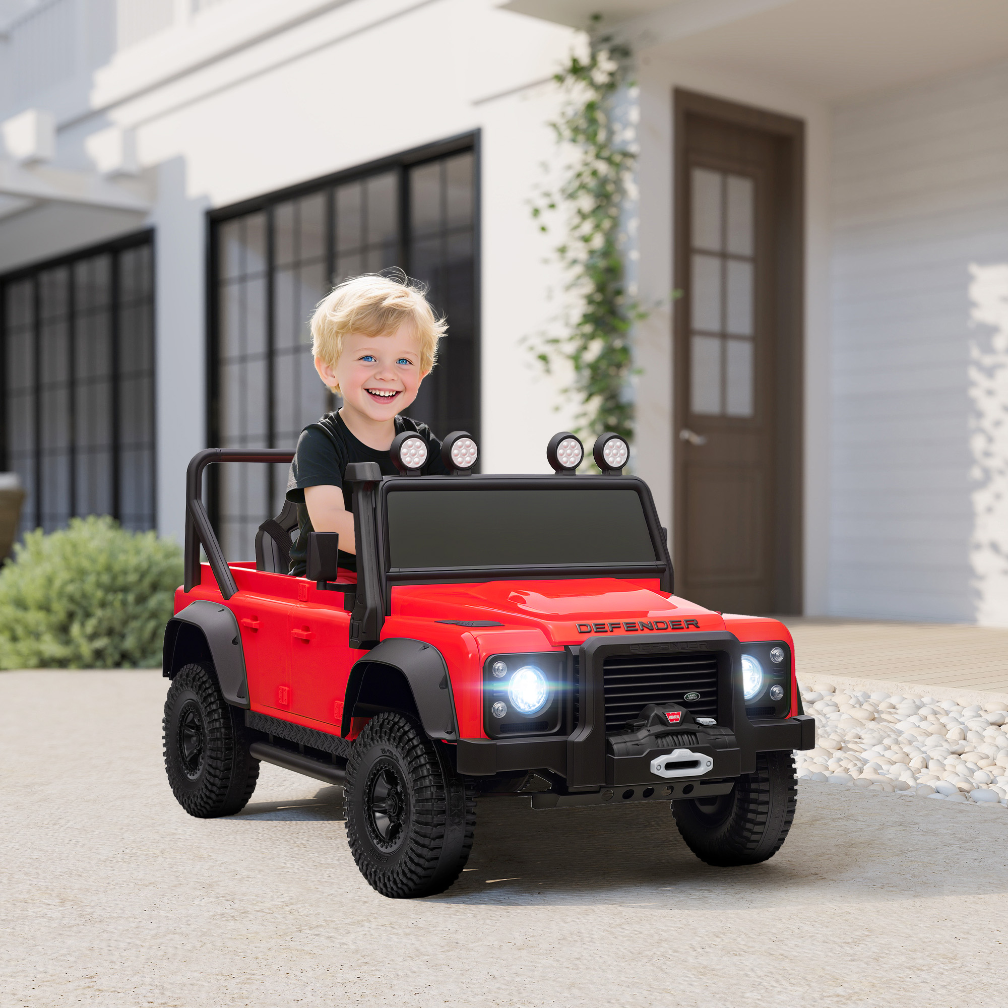 Un bambino sorridente in un'auto giocattolo rossa. L'auto è parcheggiata su cemento davanti a una casa bianca.