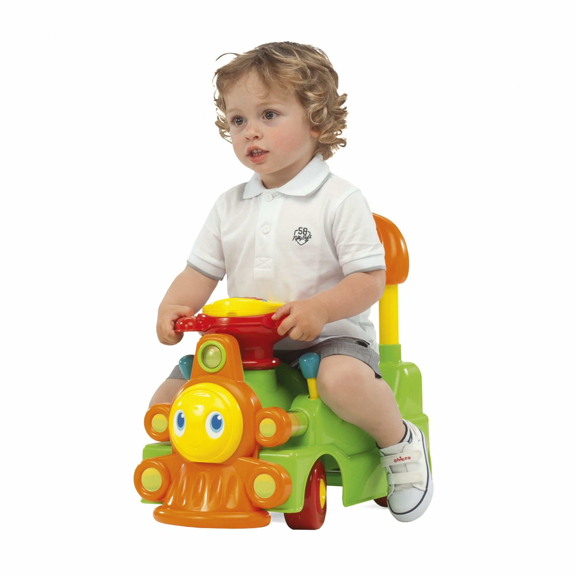 Ein kleiner Junge sitzt auf einem grünen und orangefarbenen Spielzeugzug. Er trägt ein weißes Hemd und Shorts. Der Hintergrund ist schlicht weiß.