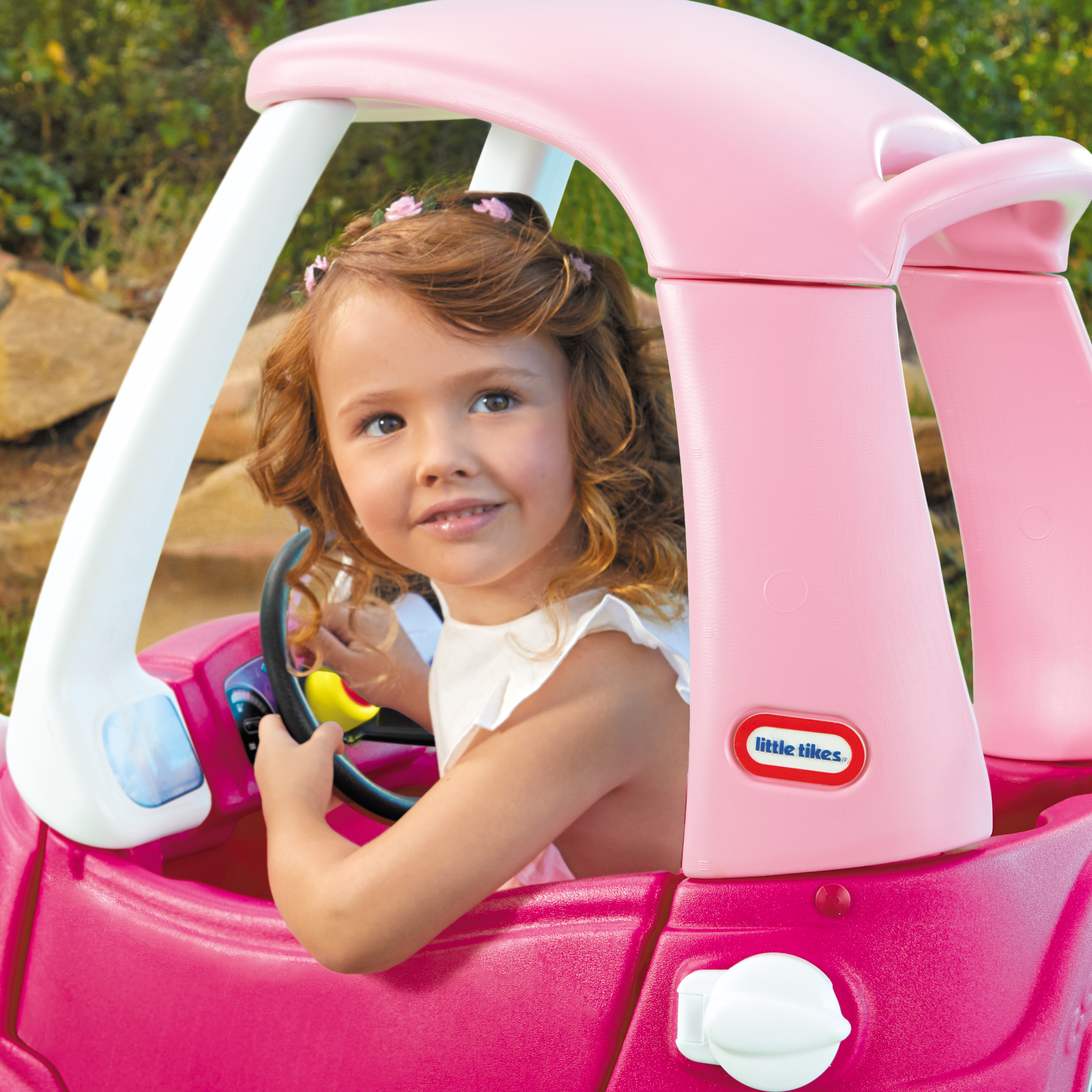 Una bambina in un'auto giocattolo rosa sorride. Ha i capelli ricci e indossa un vestito bianco. L'auto è in un giardino.