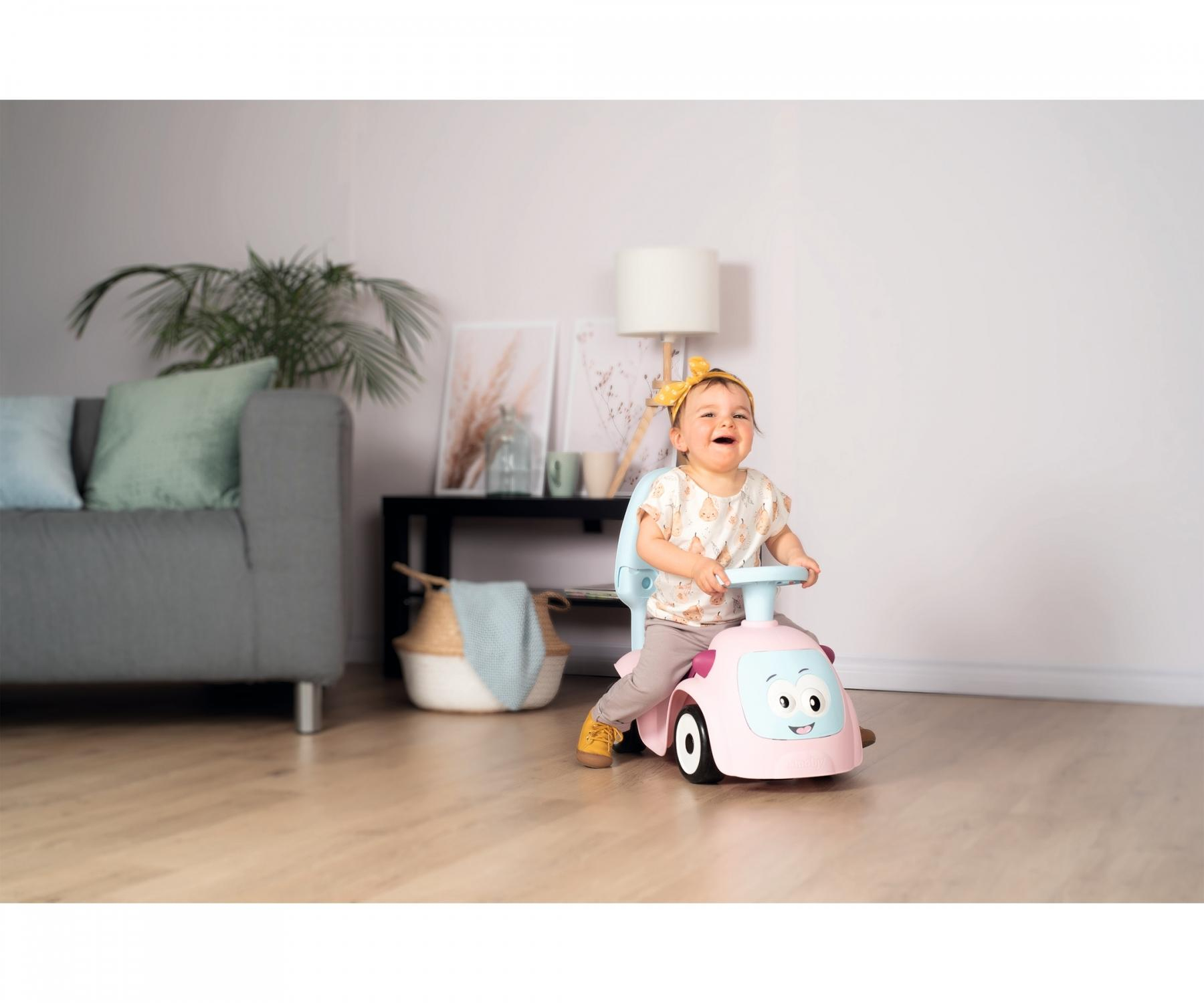 Ein lächelndes Baby sitzt in einem rosa Spielzeugauto. Das Kind trägt ein gelbes Stirnband. Eine graue Couch und Pflanzen sind im Hintergrund.