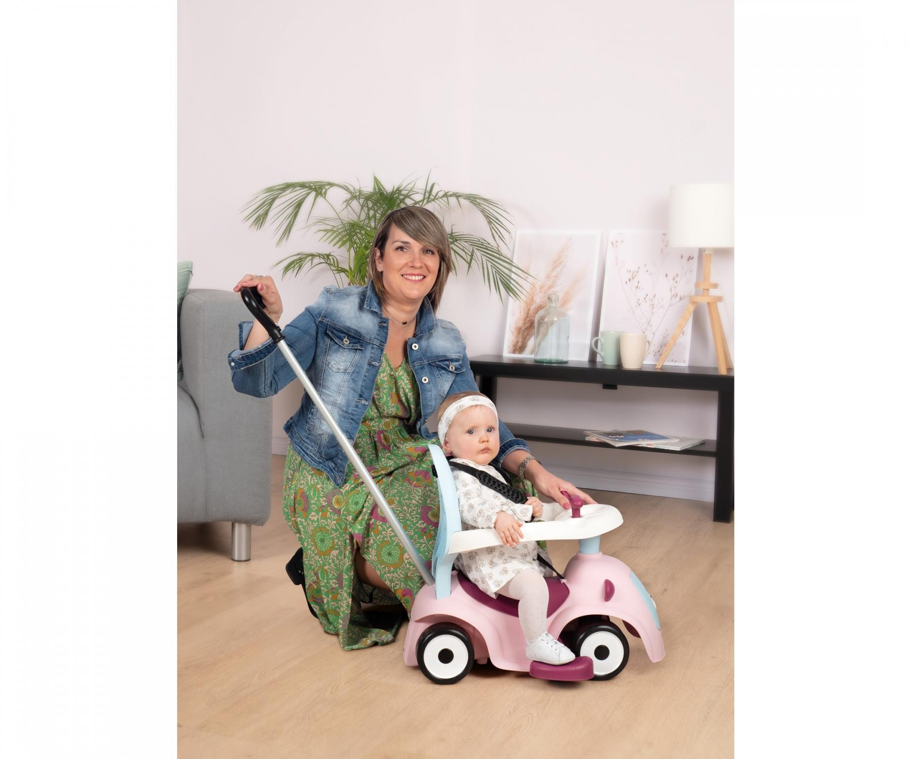 Eine Frau kniet und führt ein Baby in einem rosa Auto. Das Baby trägt ein Stirnband. Der Hintergrund umfasst eine Couch, eine Pflanze und einen Tisch.