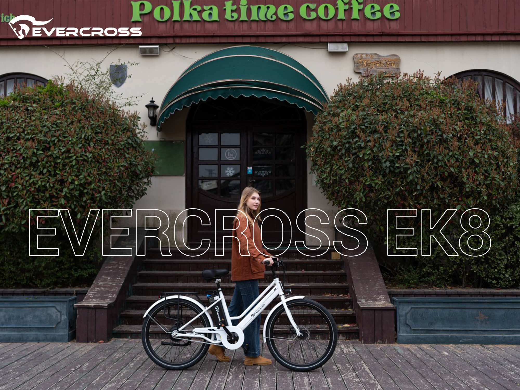 Eine Frau steht mit einem weißen Evercross EK8-Fahrrad vor einem Café mit grüner Markise. Auf dem Schild steht 'Polka time coffee'.