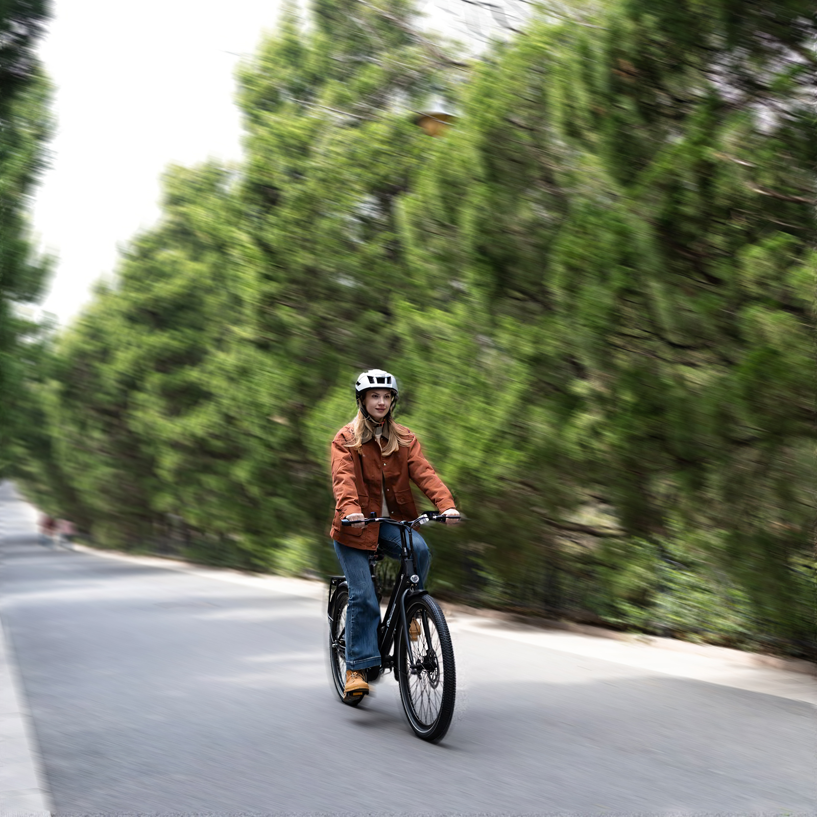 Eine Frau mit Helm fährt auf einer Straße Fahrrad, umgeben von grünen Bäumen. Sie trägt eine braune Jacke und blaue Jeans.