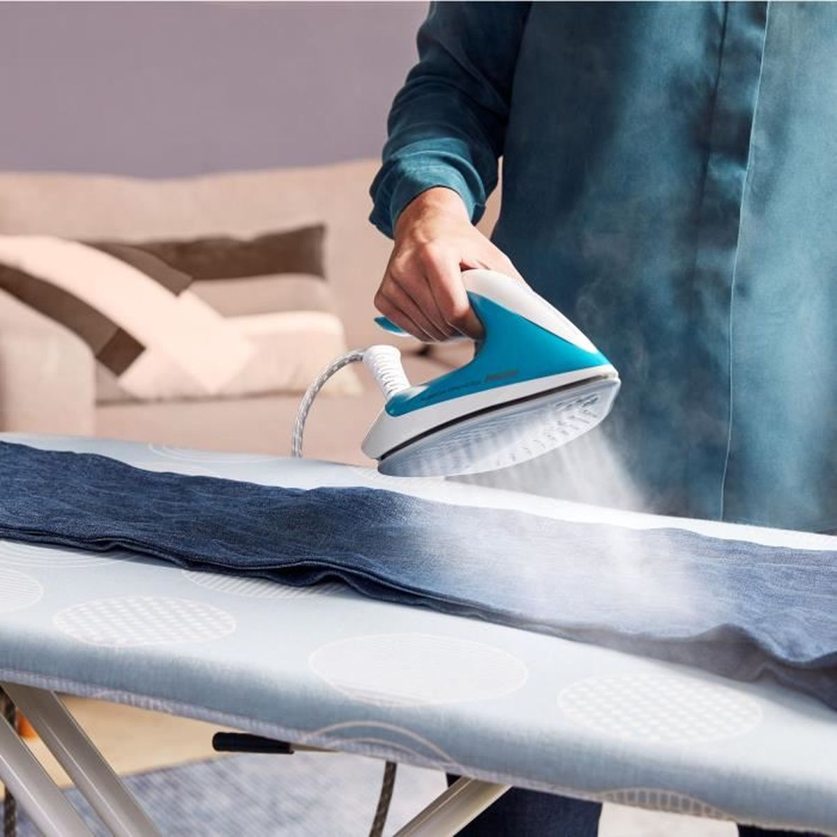 Eine Person bügelt Jeans auf einem Bügelbrett. Das Bügeleisen ist blau und weiß. Dampf tritt aus dem Bügeleisen aus.