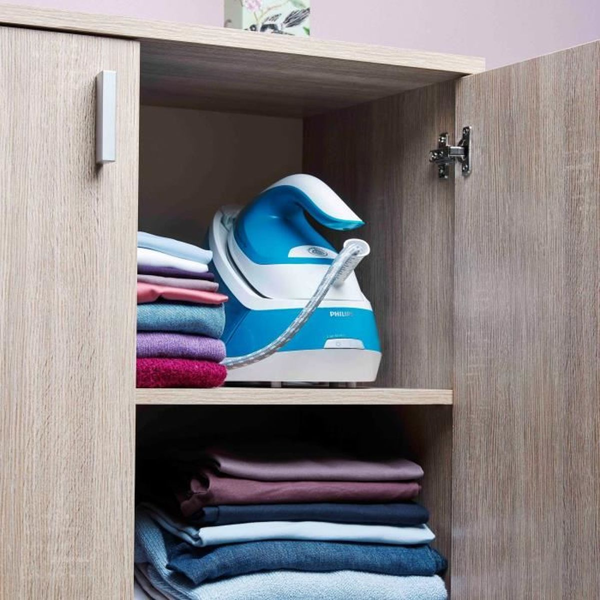 Ein blau-weißes Bügeleisen steht in einem Holzschrank mit gefalteter Kleidung. Die Schranktür ist offen und zeigt Bügeleisen und Kleidung.