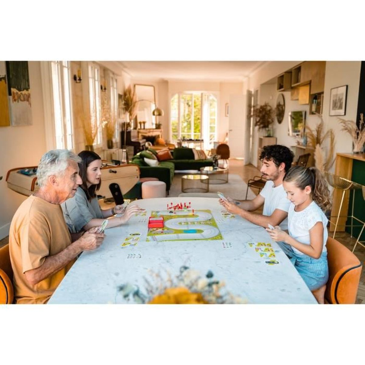 Vier mensen spelen een bordspel aan een tafel. Een man met wit haar, twee vrouwen en een man met krullend haar. Het spel staat in het midden.