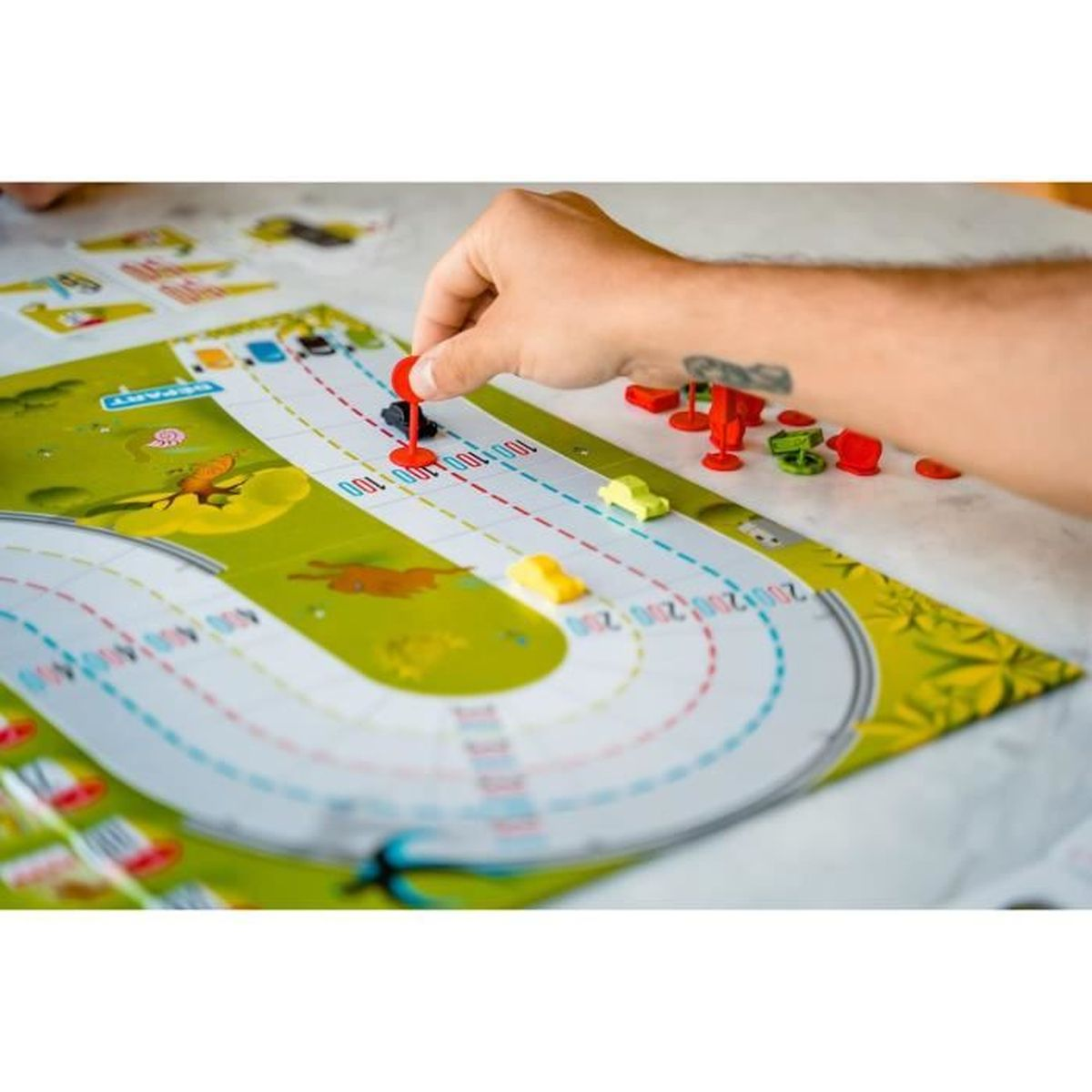 Een hand plaatst een rood spelstuk op een racebaanbord. Kleine speelgoedauto's staan op de baan. Andere spelstukken liggen in de buurt.