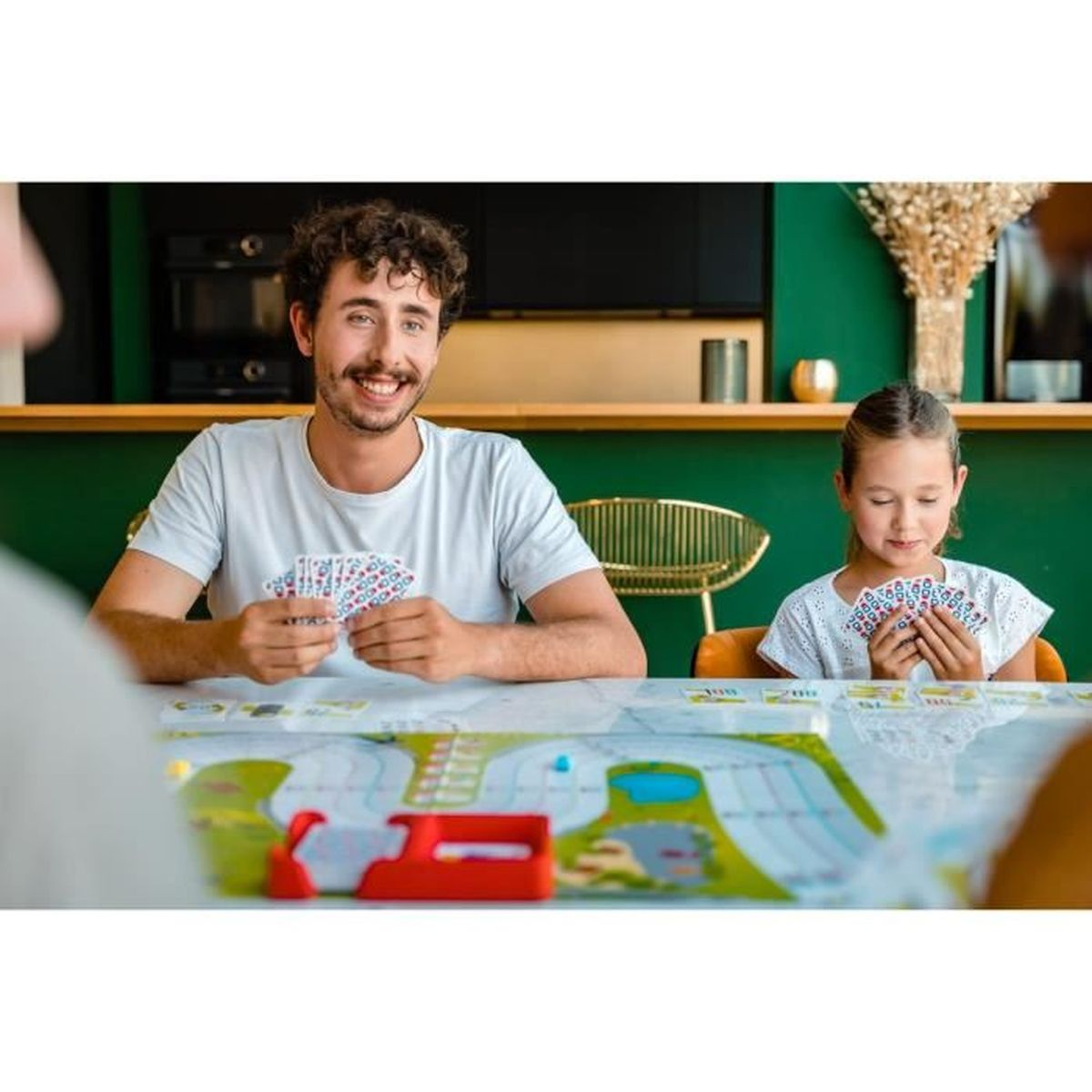 Een man en een meisje spelen kaarten. De man lacht, met kaarten in de hand. Het meisje kijkt naar haar kaarten. Er ligt een spelbord op tafel.