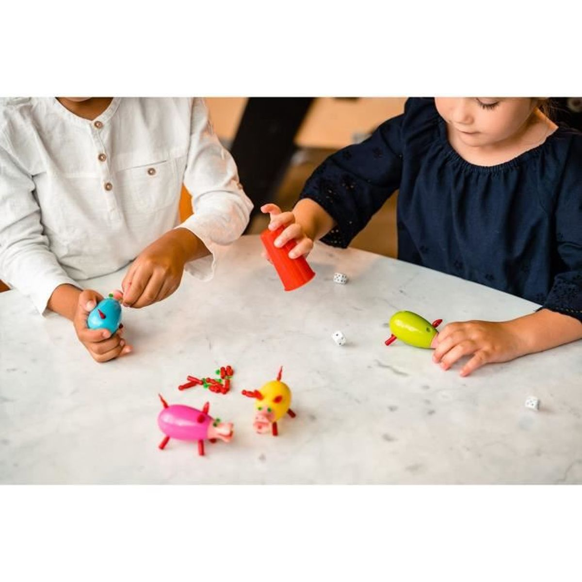 Bambini che giocano con maialini giocattolo, dadi e una tazza rossa su un tavolo di marmo. Sono presenti due bambini. Il gioco sembra un'attività divertente.