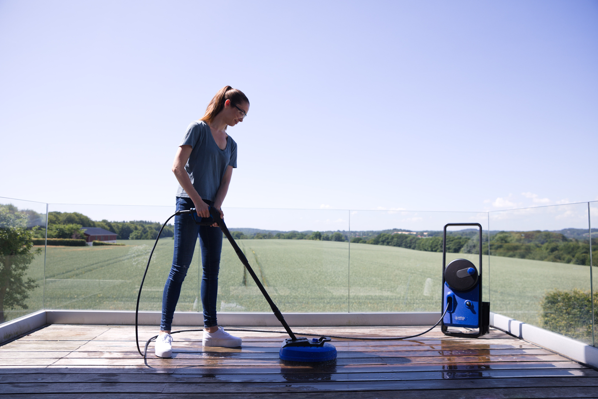 Kobieta czyści drewniany taras myjką ciśnieniową. Zielone pola i drzewa w tle. Niebo jest czyste i niebieskie.