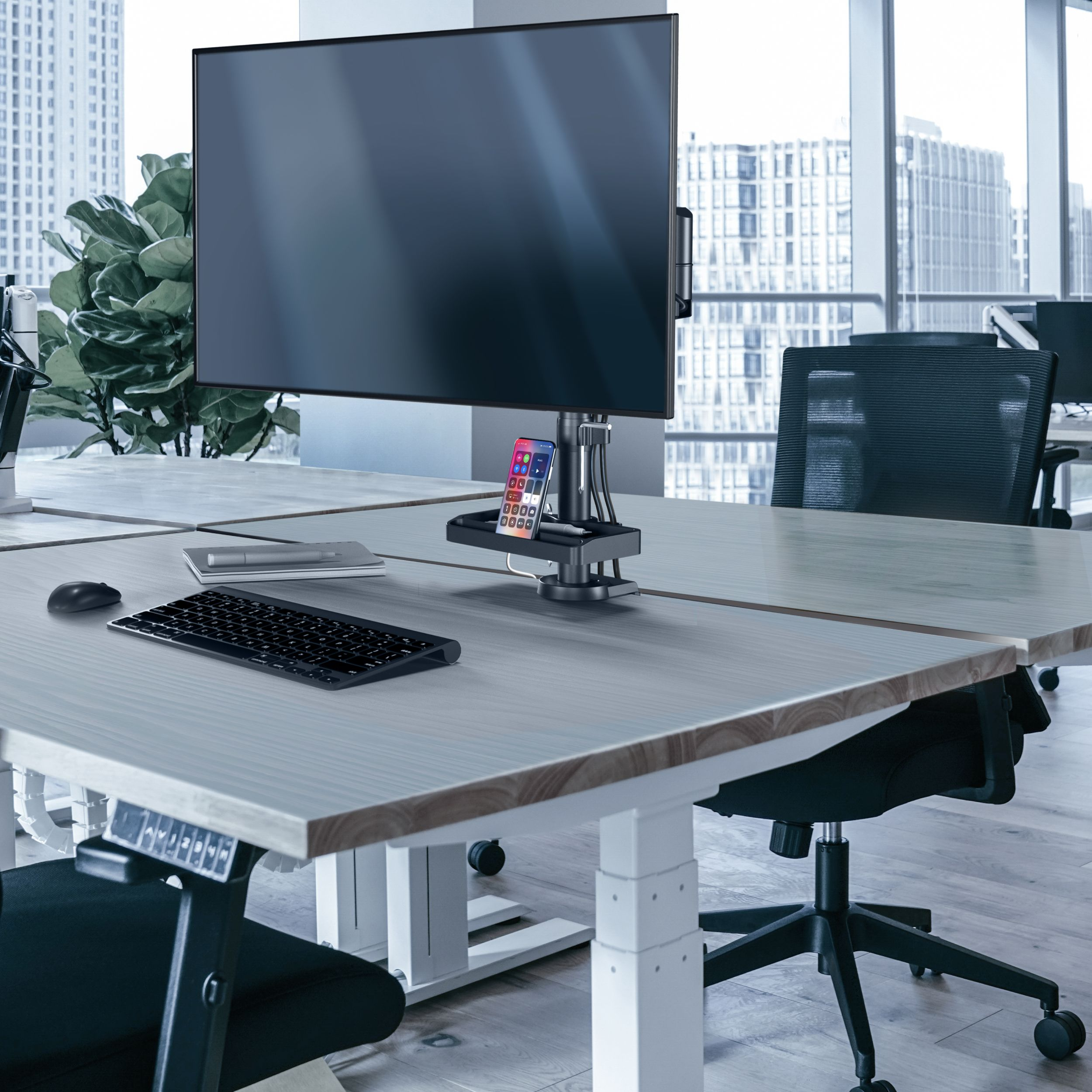 Scrivania con monitor, tastiera, mouse e smartphone su supporto. Sedia sullo sfondo. Vista sulla città.