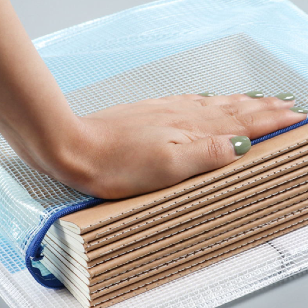 Dłoń z zielonymi paznokciami spoczywa na stosie notatników w siatkowej torbie. Niebieski zamek błyskawiczny biegnie wzdłuż górnej krawędzi torby.