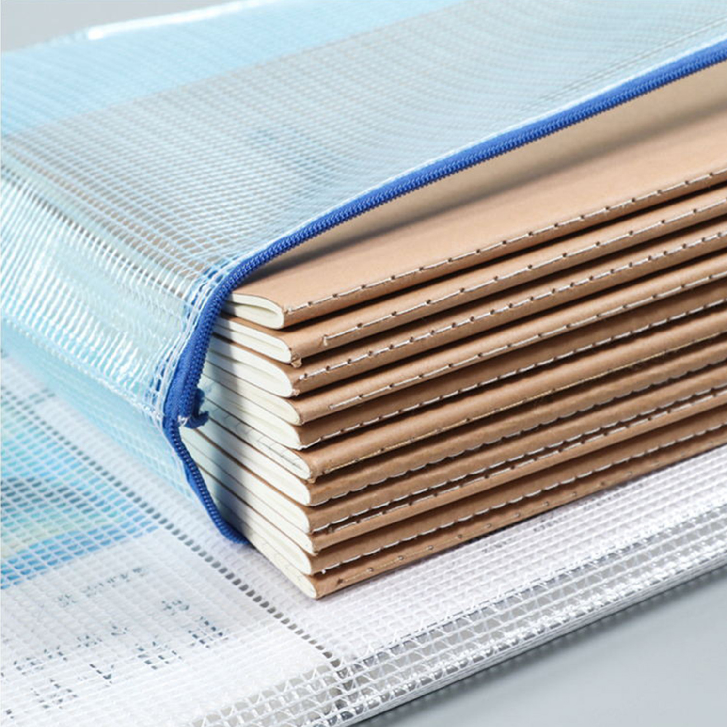 Stos notatników w niebieskiej siatkowej torbie zapinanej na zamek. Widoczne brązowe okładki. Zamek błyskawiczny jest niebieski. Tło jest szare.