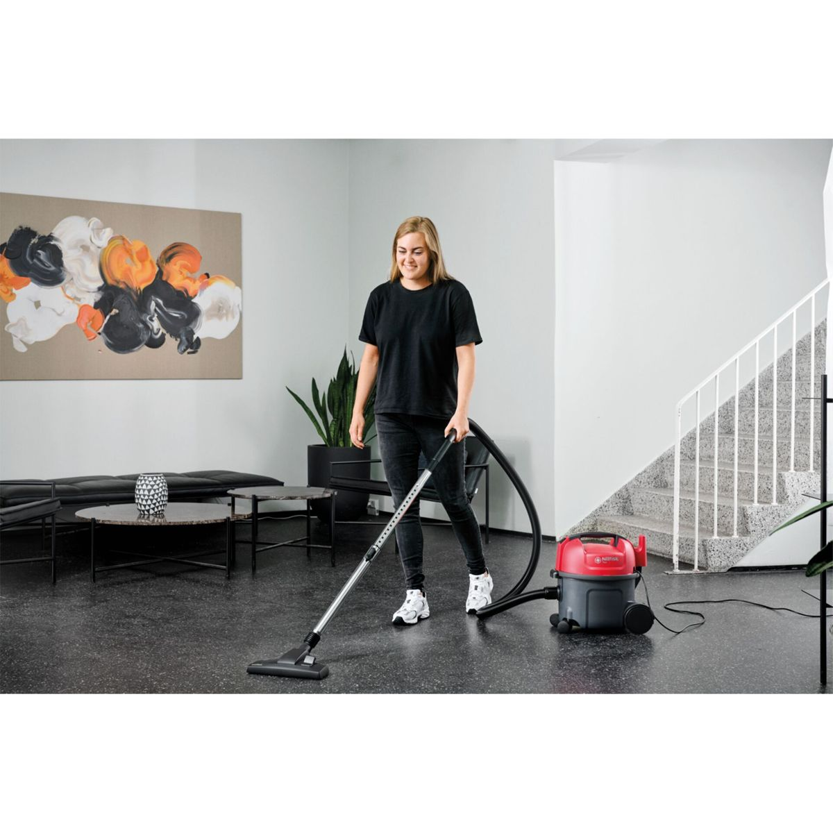 Kobieta odkurza pokój. Ma na sobie czarną koszulkę i dżinsy, używając odkurzacza. Widoczny jest czerwono-czarny odkurzacz.