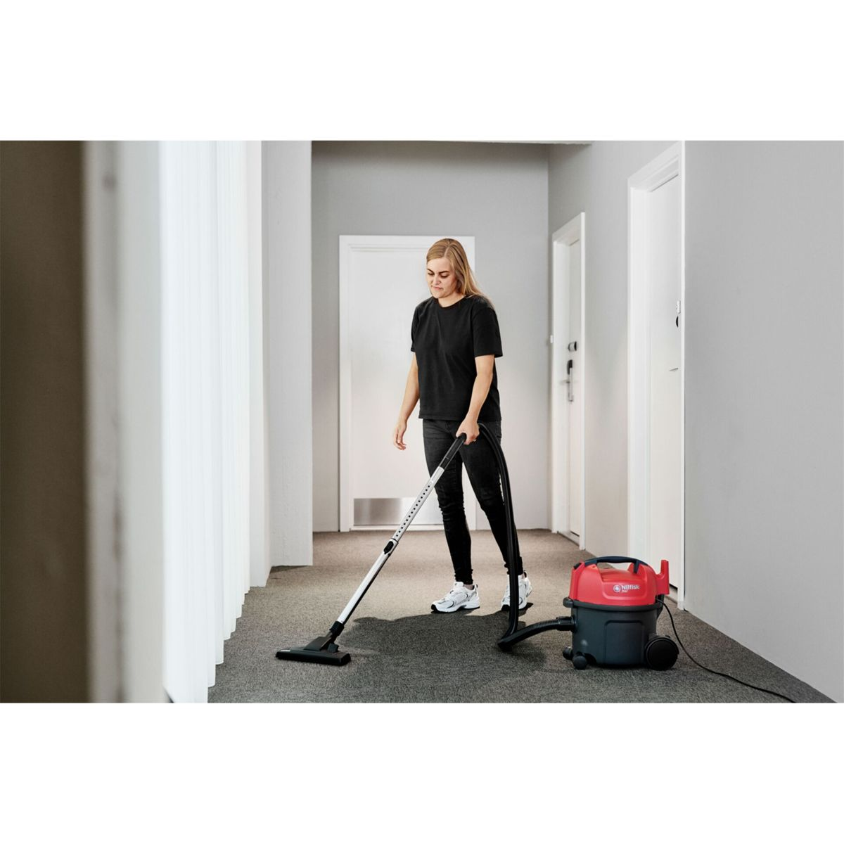 Kobieta odkurza wykładzinę na korytarzu. Ma na sobie czarne ubrania i białe trampki. Obok niej stoi czerwono-czarny odkurzacz.