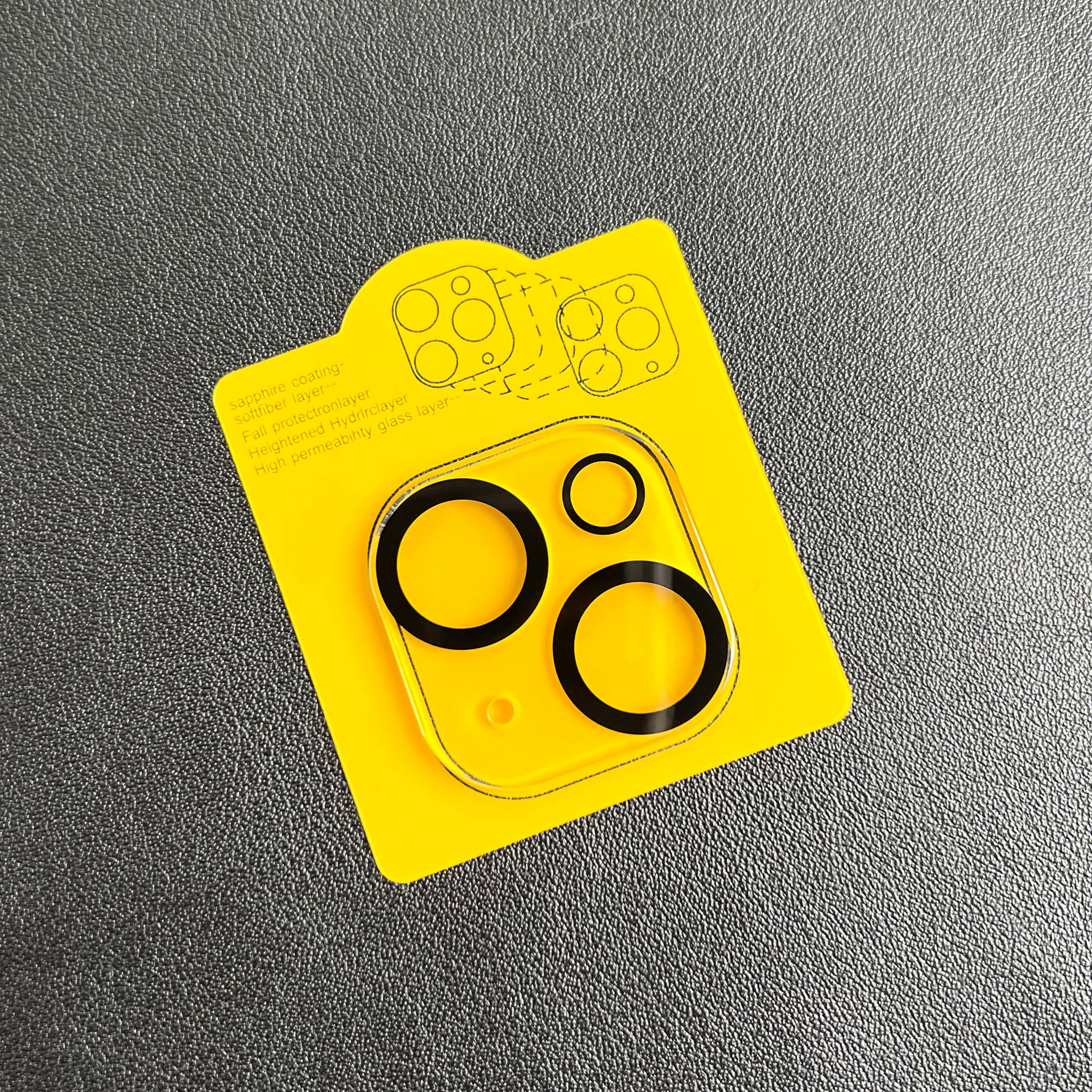 Een gele kaart met een lensbeschermer voor de telefooncamera. De beschermer is transparant met zwarte ringen op een grijs oppervlak.