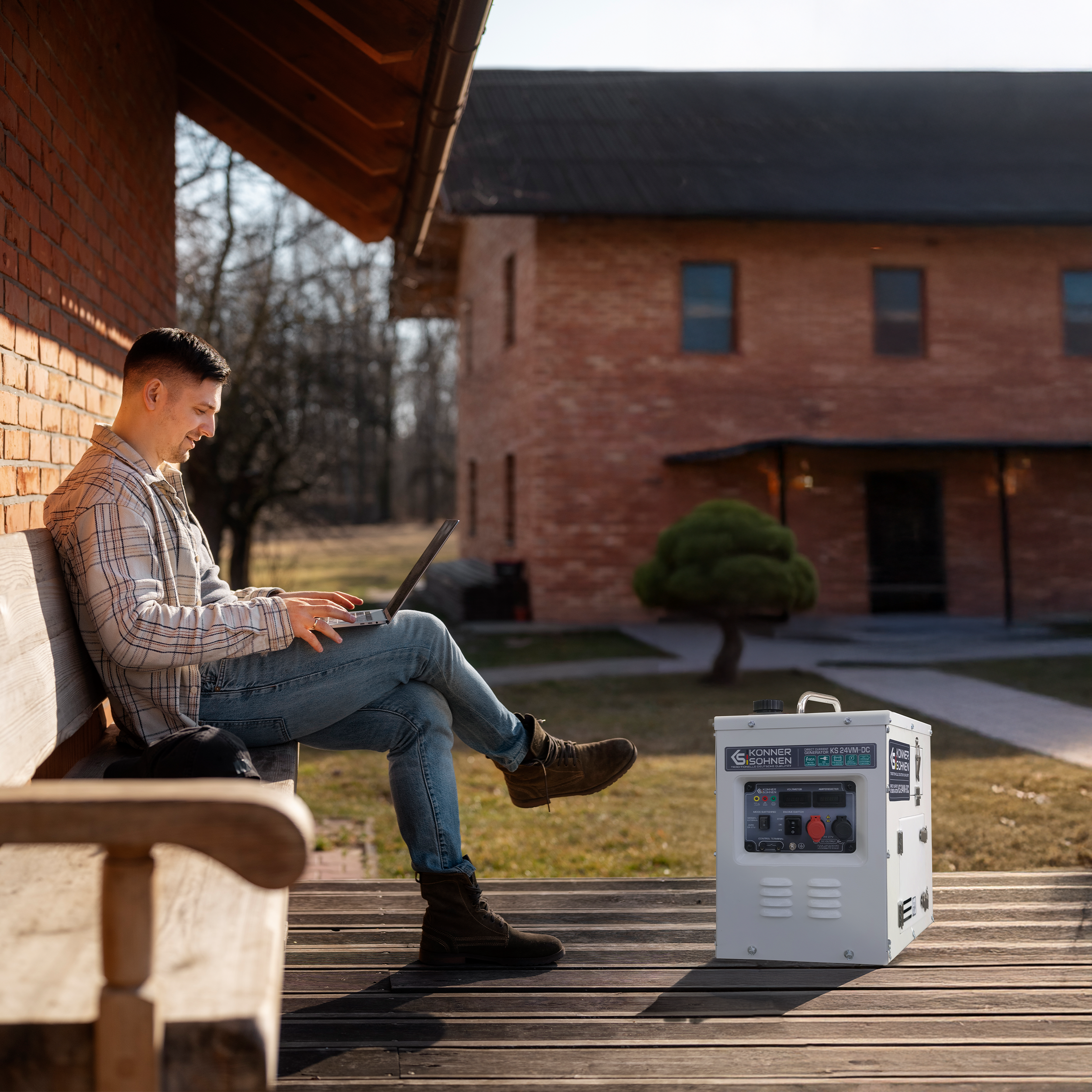 Mężczyzna siedzi na ławce, używając laptopa. W pobliżu stoi generator. W tle ceglany budynek.